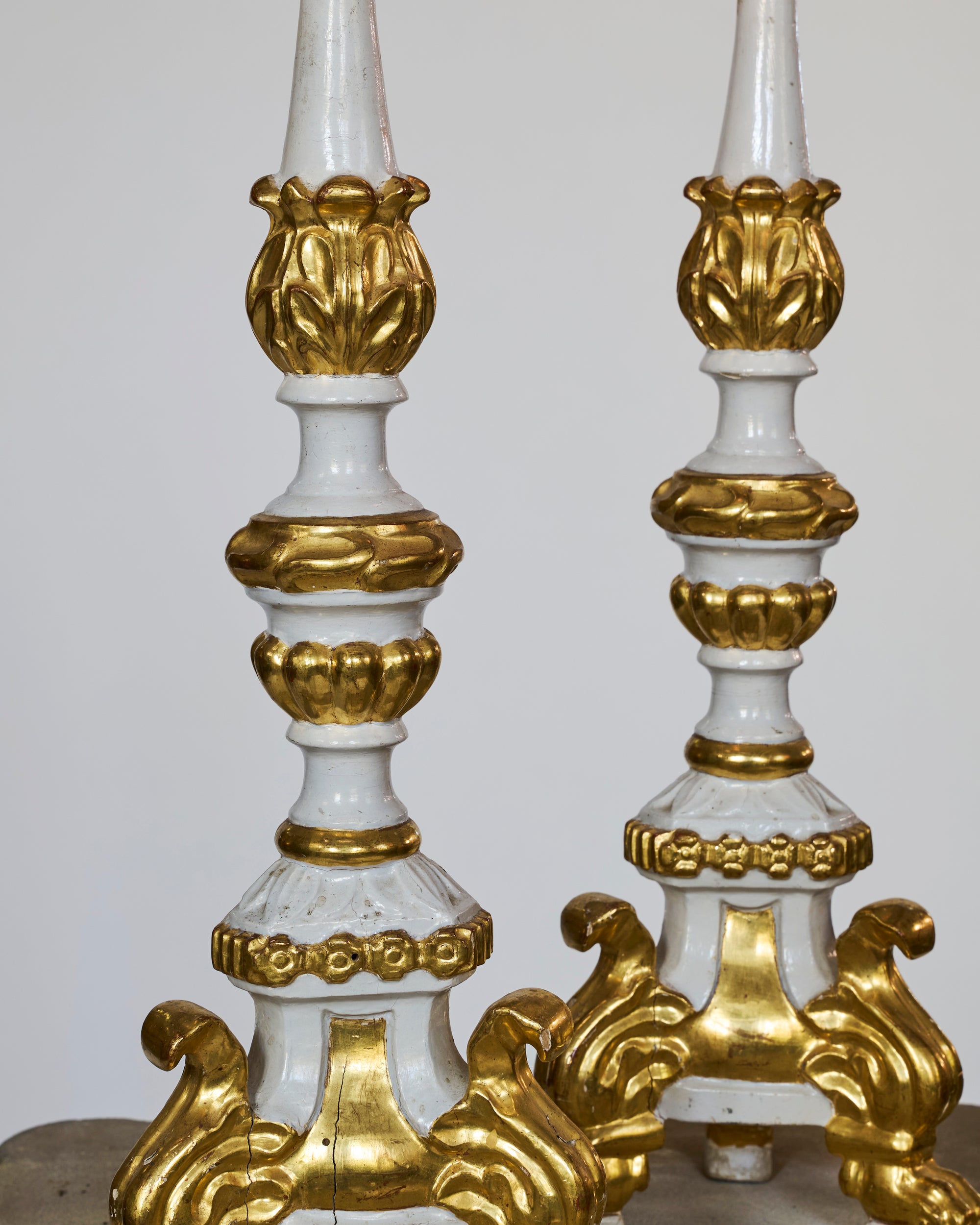 A pair of Antique European Giltwood Candlesticks with a white painted finish stand side by side, showcasing intricate carved details and decorative bases against a plain background.