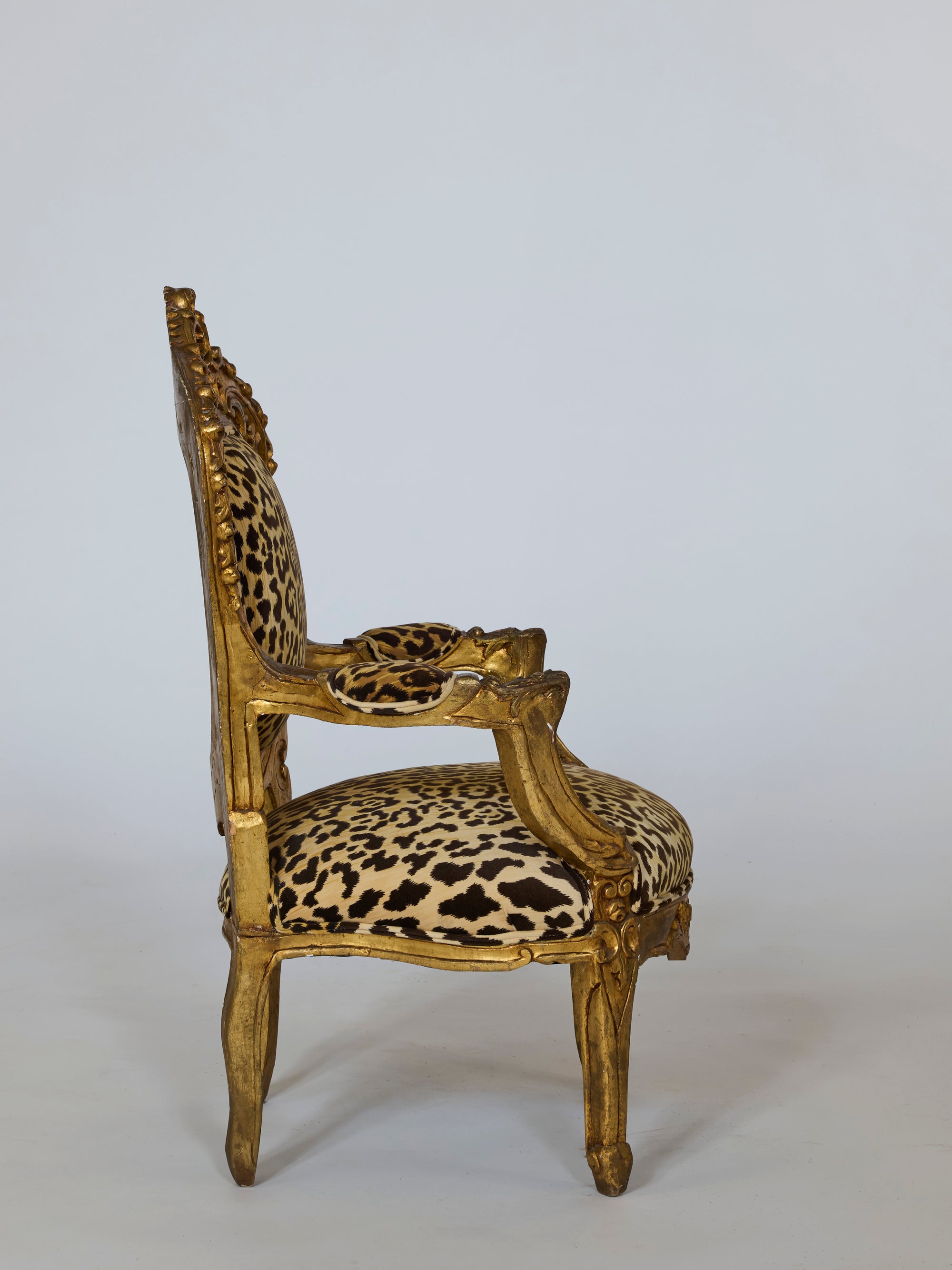A side view of a Pair of Leopard Upholstered Gilt Armchairs displays ornate, gold-painted Baroque frames with intricate carvings and luxurious leopard-print fabric on the seat, back, and arms, set against a plain light background.