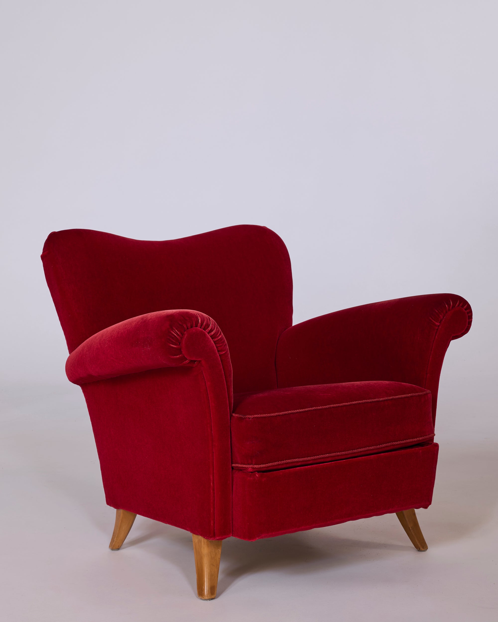A pair of red velvet club armchairs with sculpted rolled arms and wooden legs is set against a plain light background, evoking the classic vintage European lounge chair style.