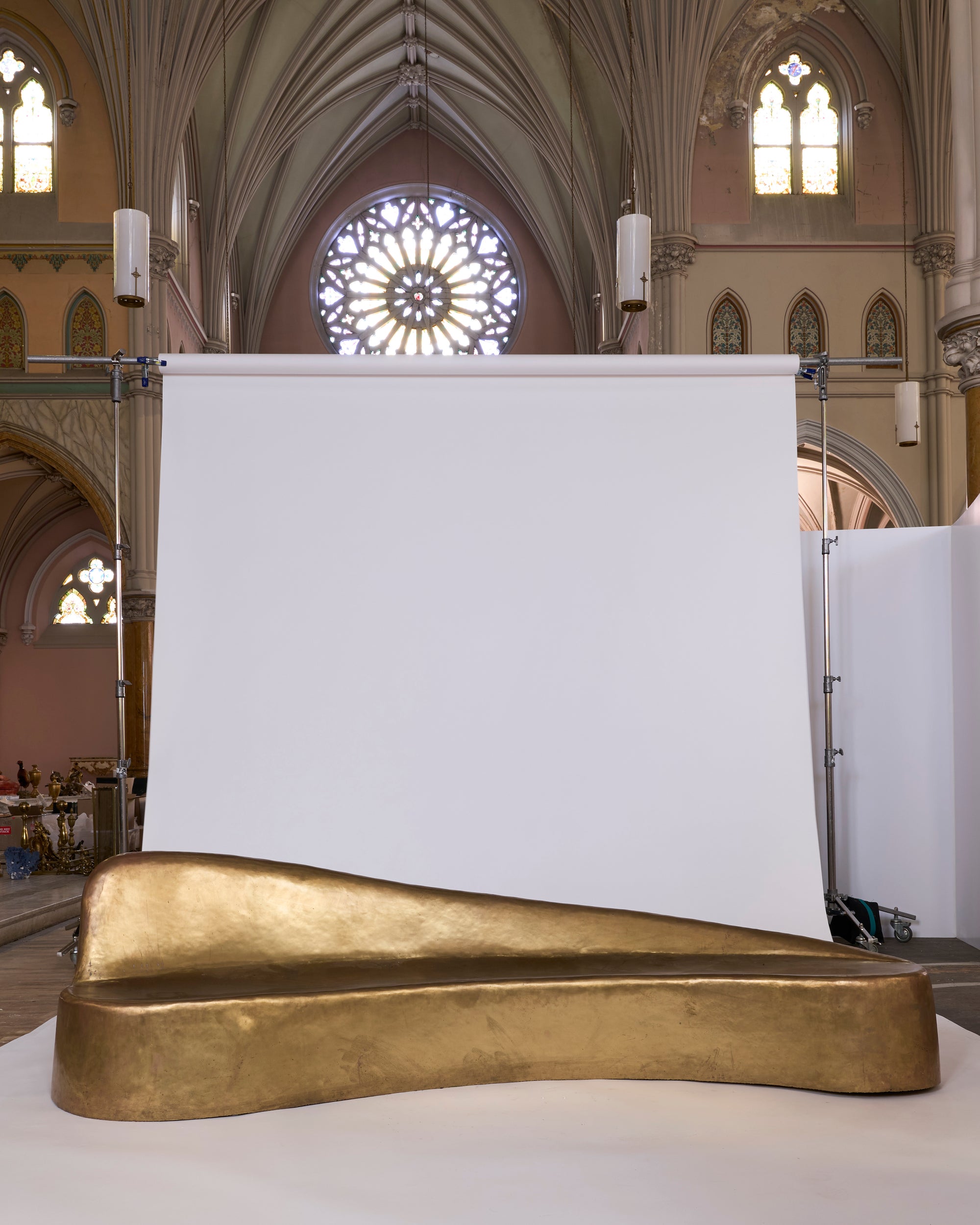The Sculptural Brass Chaise Lounge with Organic Curved Form stands before a white backdrop in a cathedral, surrounded by tall arches, stained glass windows, and ornate architectural details.