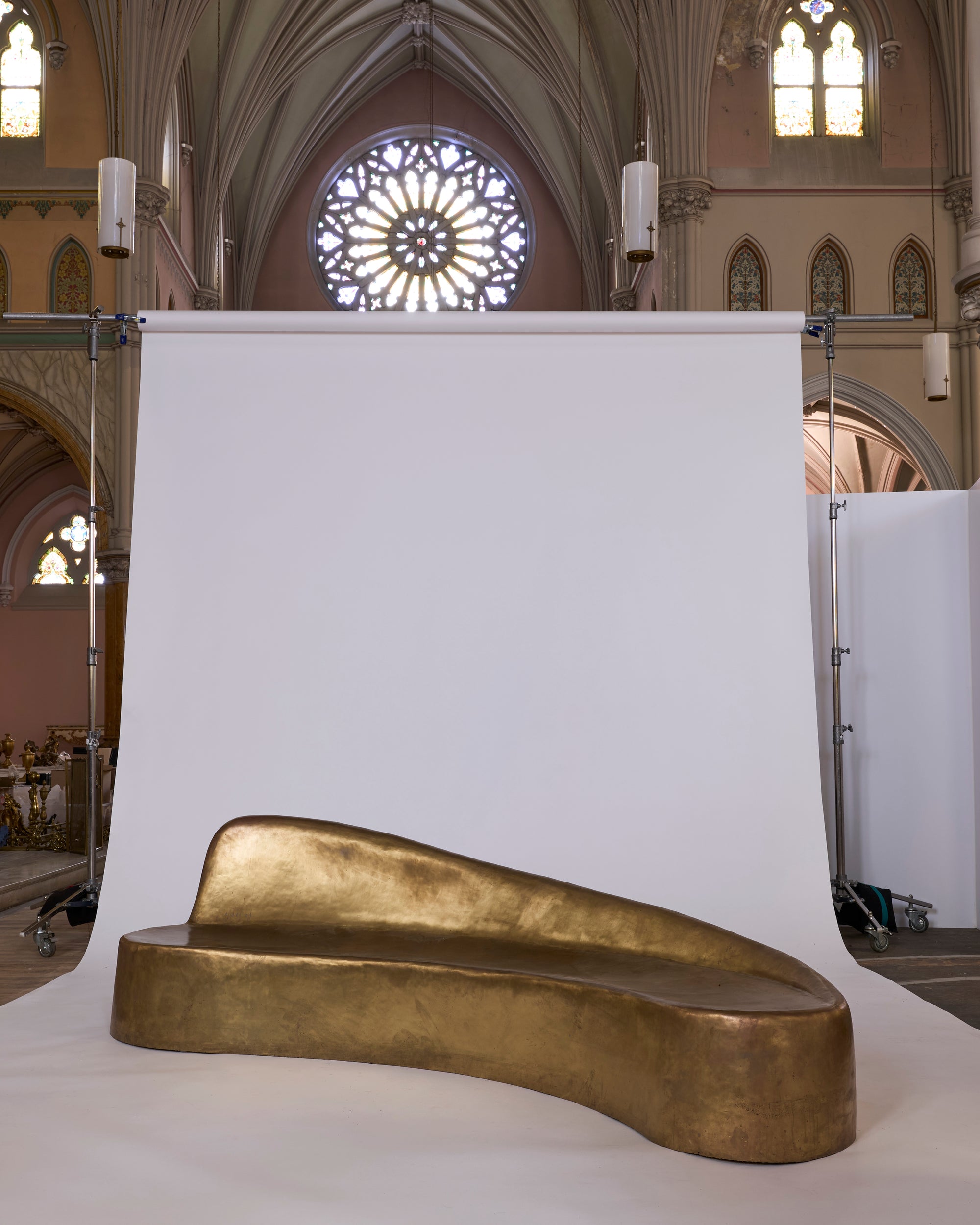 The Sculptural Brass Chaise Lounge with Organic Curved Form is positioned before a white backdrop inside a church featuring arched ceilings, ornate windows, and stained glass.