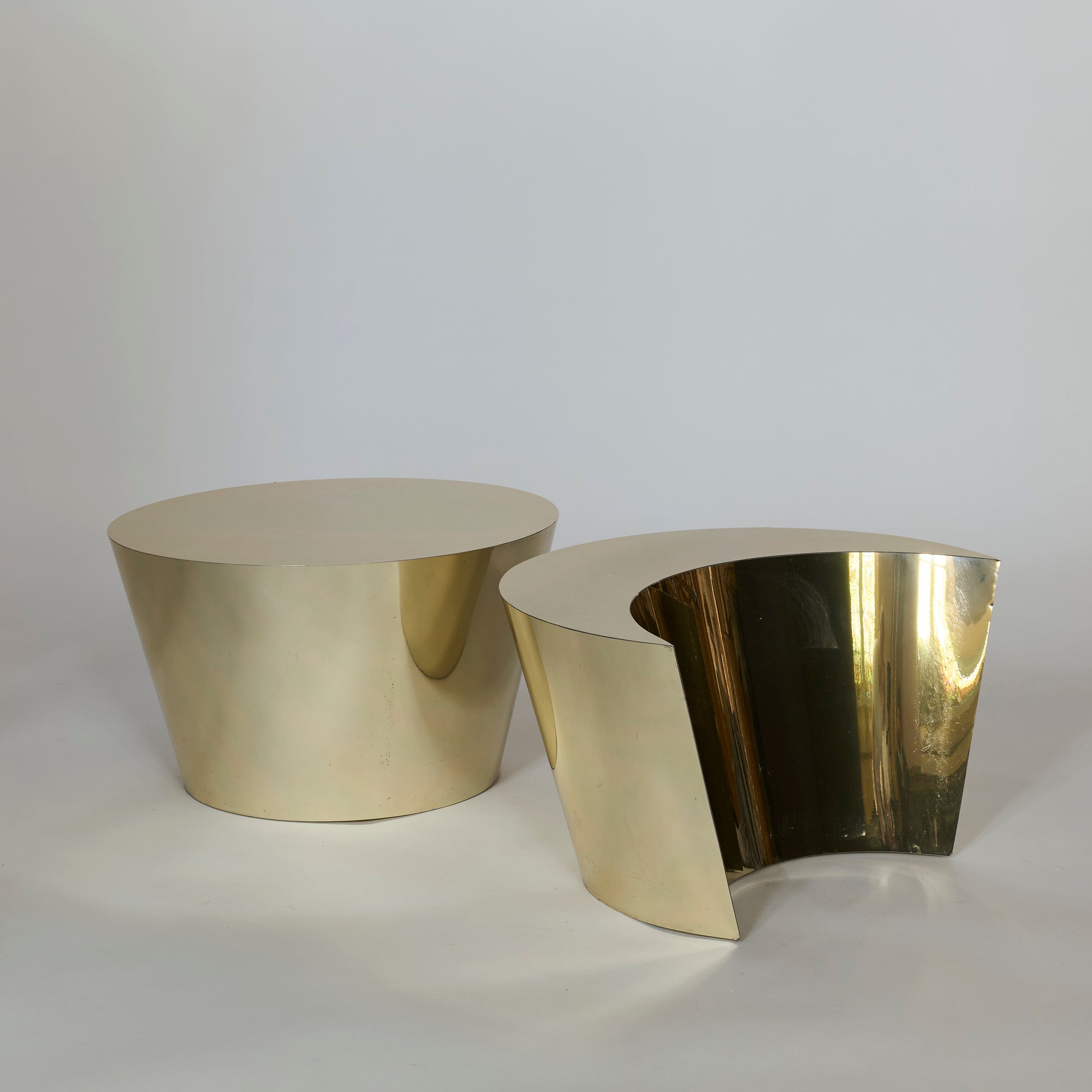 The Laminate Gold Table Set features two modern, metallic tables—one round with smooth sides and one with a sculptural wedge cutout—both boasting reflective surfaces and a sleek, minimalist design for contemporary spaces.