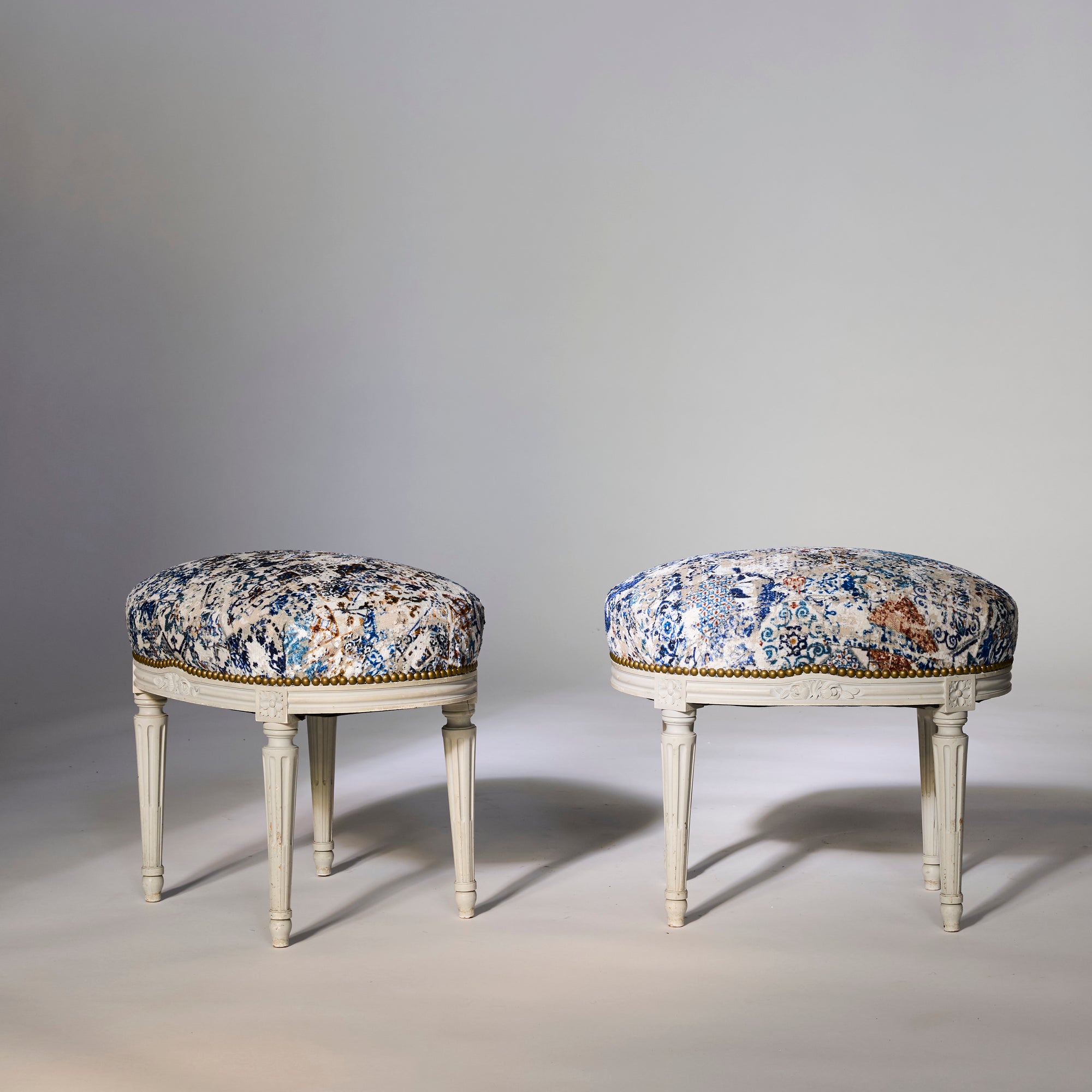 A pair of painted Louis-style stools with carved legs and distressed blue and cream upholstery showcase European elegance, sitting side by side on a light surface against a plain background.