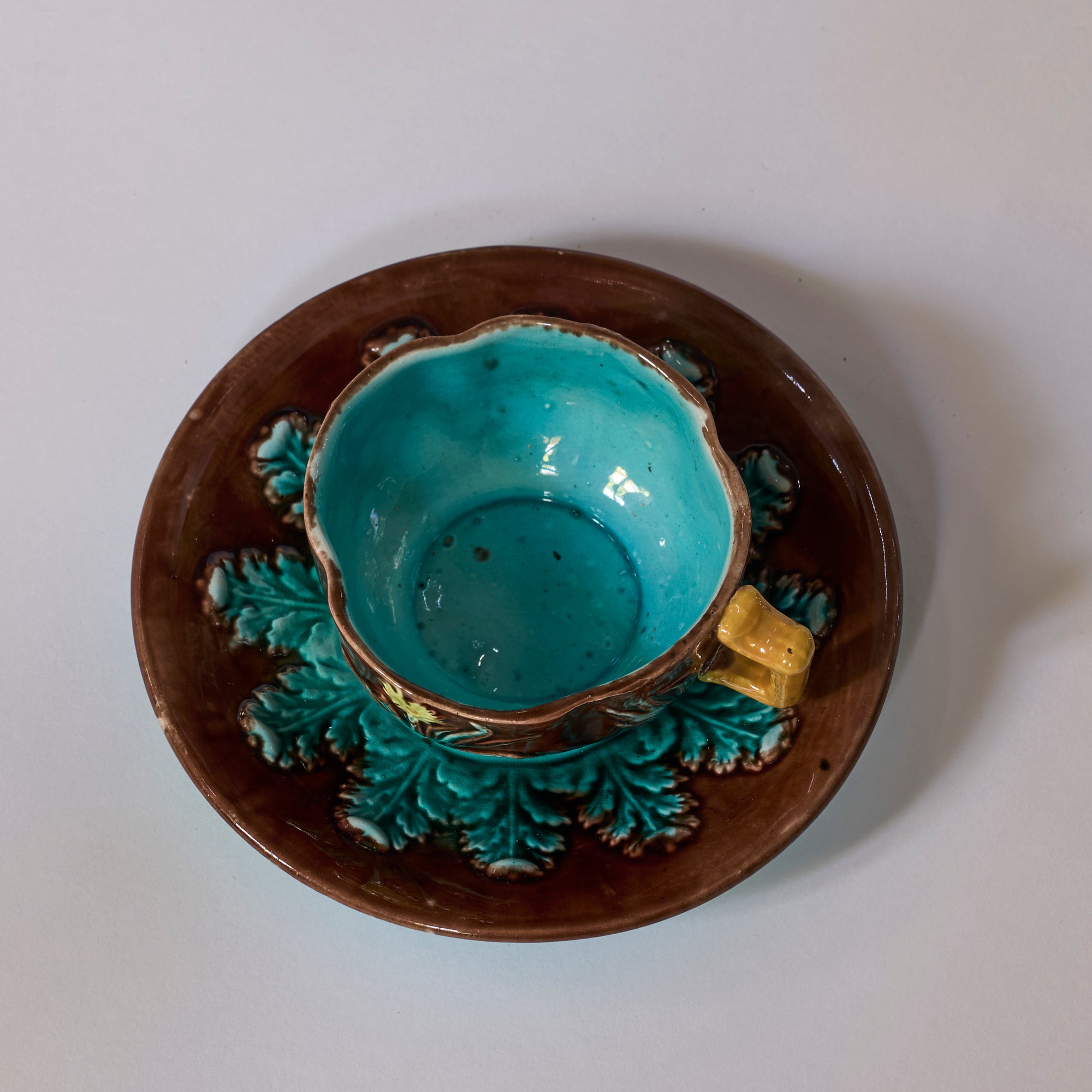 The Majolica Floral Teacup and Saucer with Turquoise Glaze features teal and brown leaf designs, a golden handle, and a bright turquoise-glazed interior, creating a striking decorative art ceramics set.