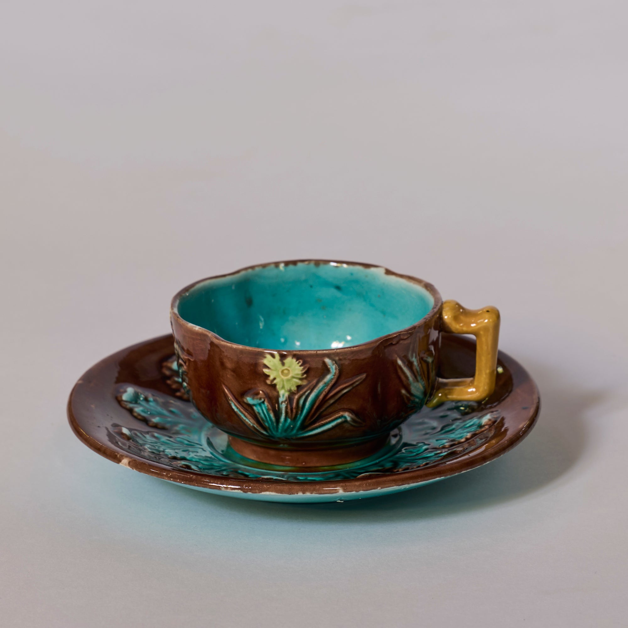 The Majolica Floral Teacup and Saucer with Turquoise Glaze features a glossy brown exterior, yellow handle, embossed floral patterns, and a vibrant turquoise interior—perfect for decorative art ceramics enthusiasts.