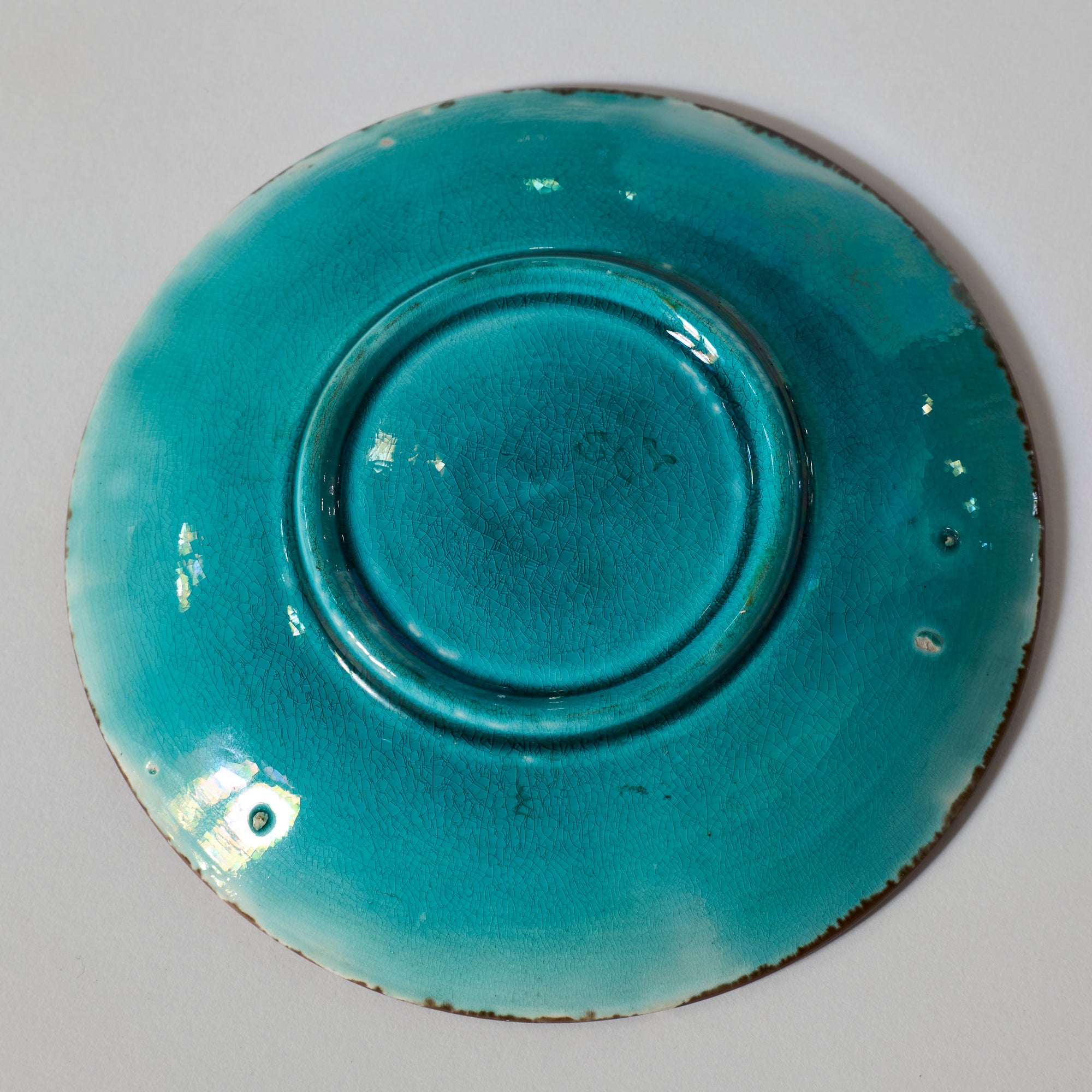 The Majolica Floral Teacup and Saucer with Turquoise Glaze features a circular raised base and subtle glaze cracks, reminiscent of decorative ceramics, shown from below against a plain white background.