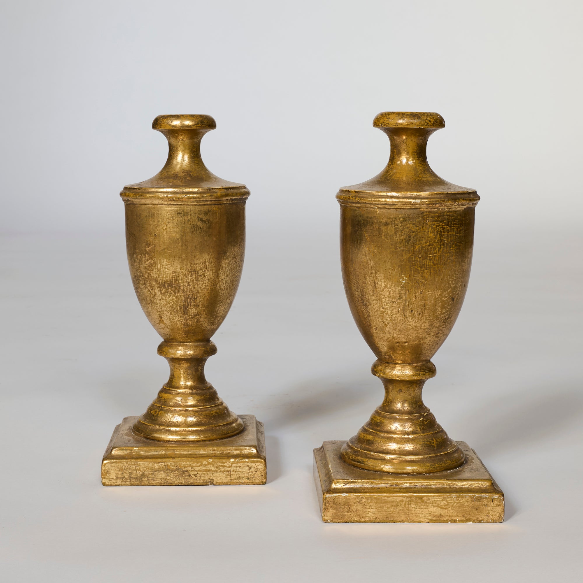A Pair of Antique Giltwood Decorative Urns with round bases and square plinths stand side by side on a light background, bringing classic charm as elegant gold-colored accents.