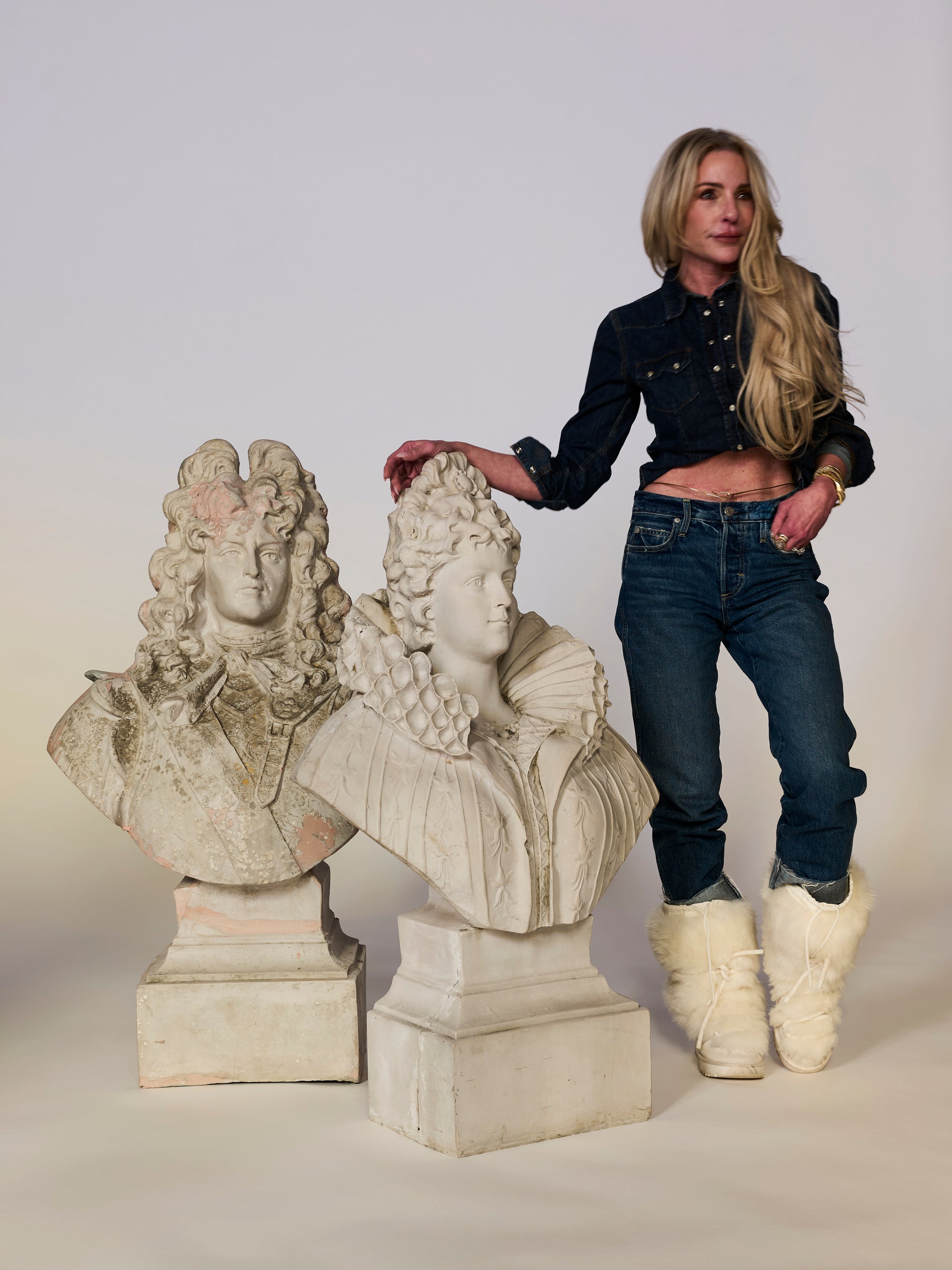 Woman standing next to two classical busts on a plain background