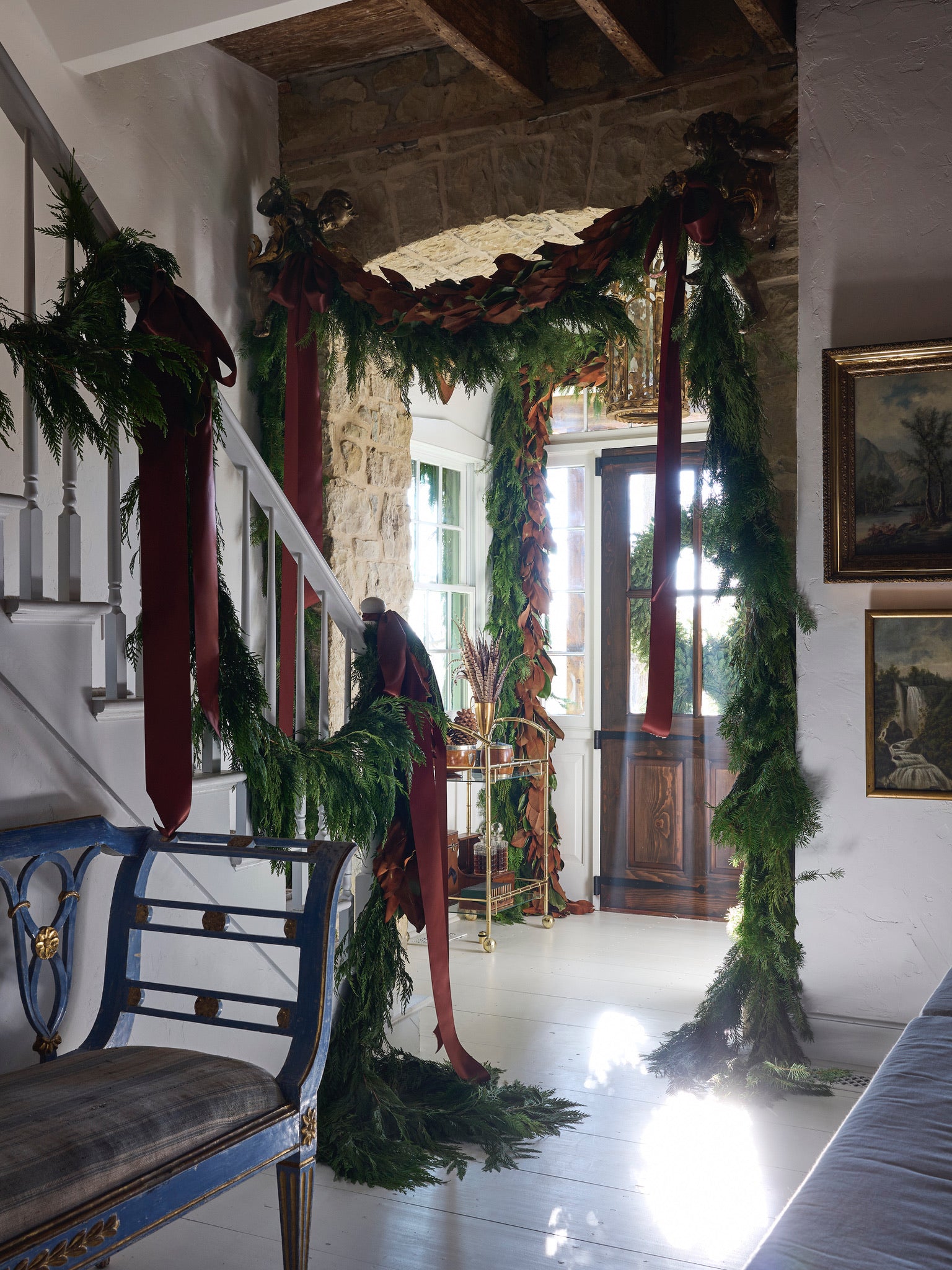 A rustic entryway decorated for the holidays with lush green garlands and large burgundy ribbons draped along the staircase and doorway, wooden beams above, and natural light streaming through a glass door.