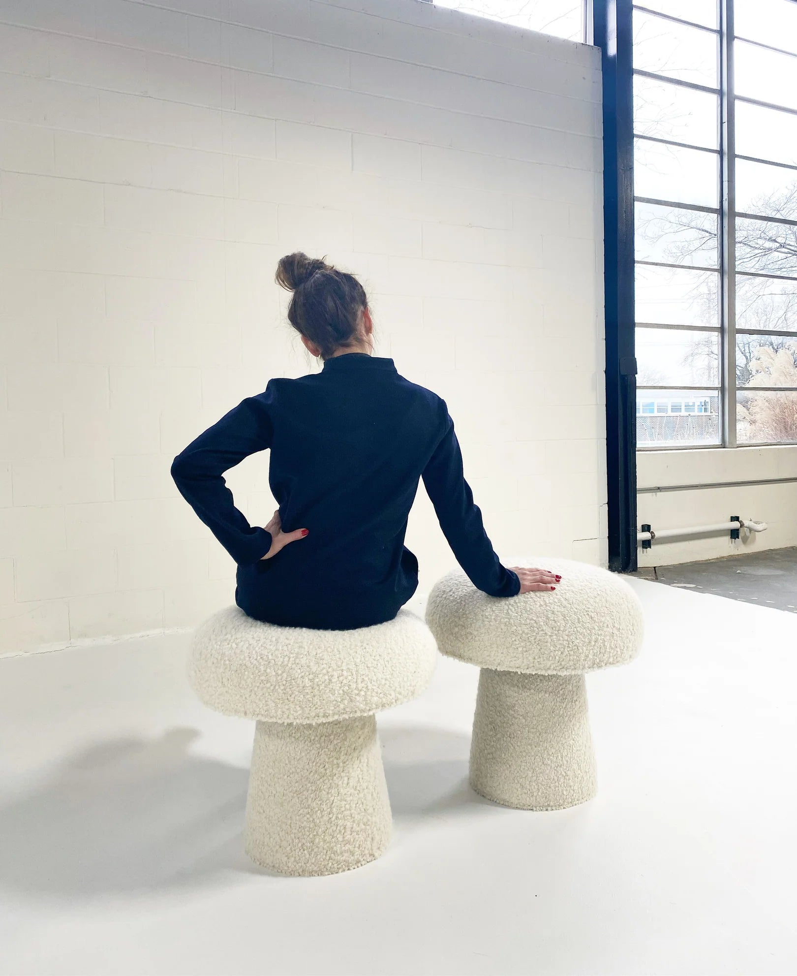 A person with hair in a bun, dressed in dark clothing, sits on The Forsyth Mushroom Pouf in Boucle—a round, textured white stool—beside a similar pouf in a bright modern room with large windows.