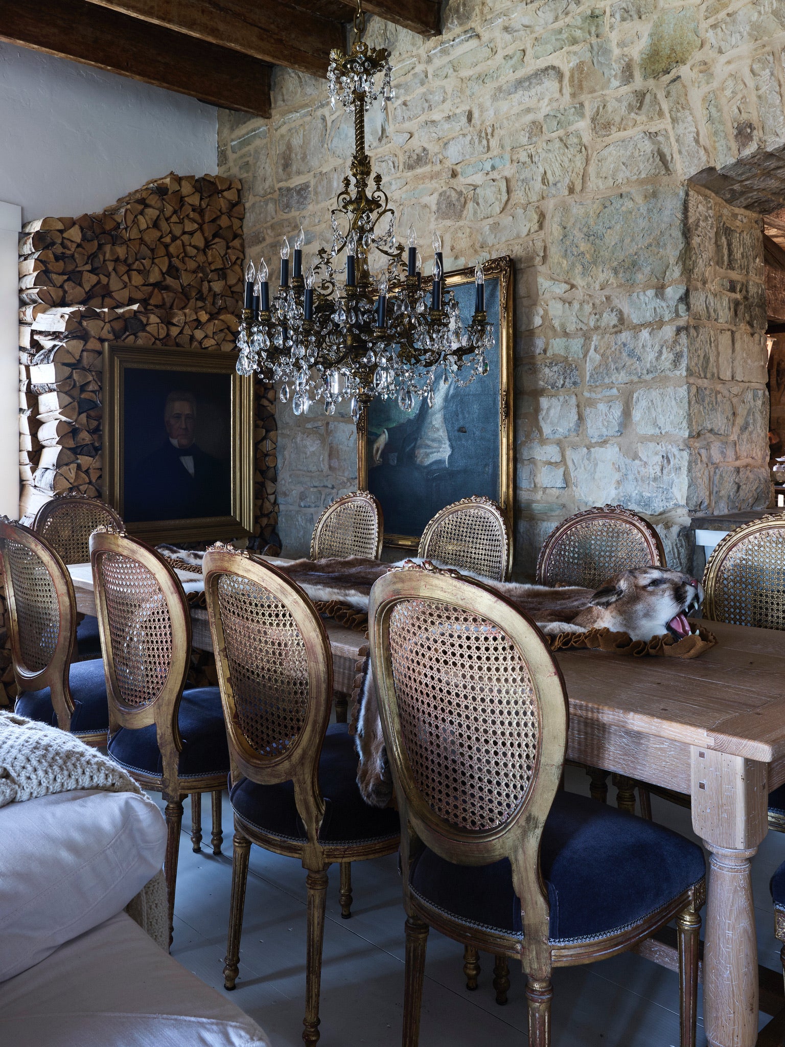 A rustic dining room with a long wooden table, vintage upholstered chairs, a crystal chandelier, stone walls, stacked firewood, and two framed portraits. The space has a cozy and historic atmosphere.