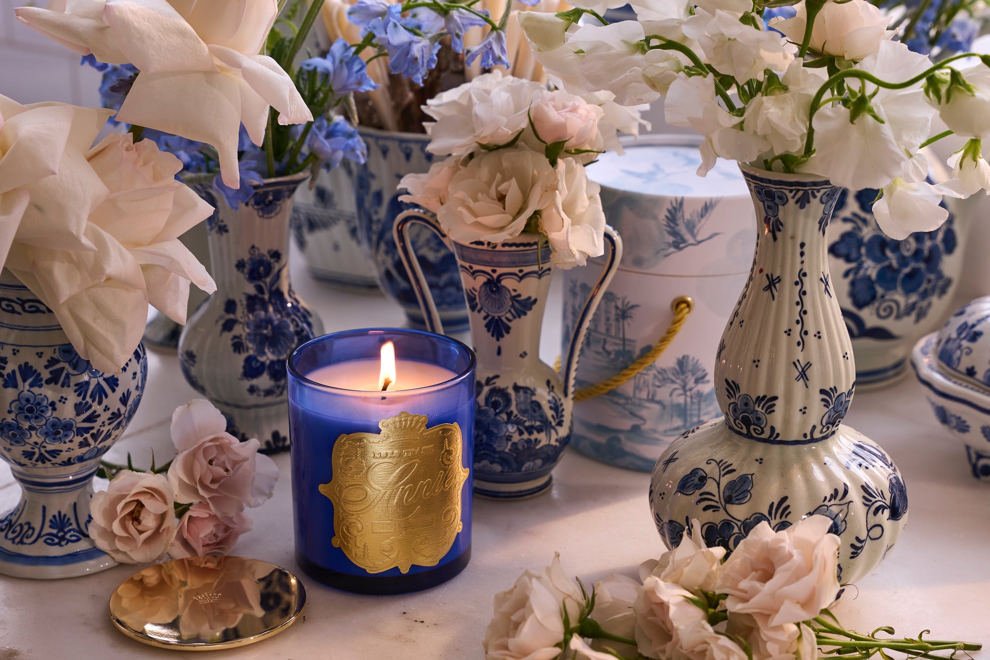 The Holland blue scented soy candle burns on a table with blue and white porcelain vases holding white and pale pink flowers; some blossoms and a gold compact mirror are scattered nearby.