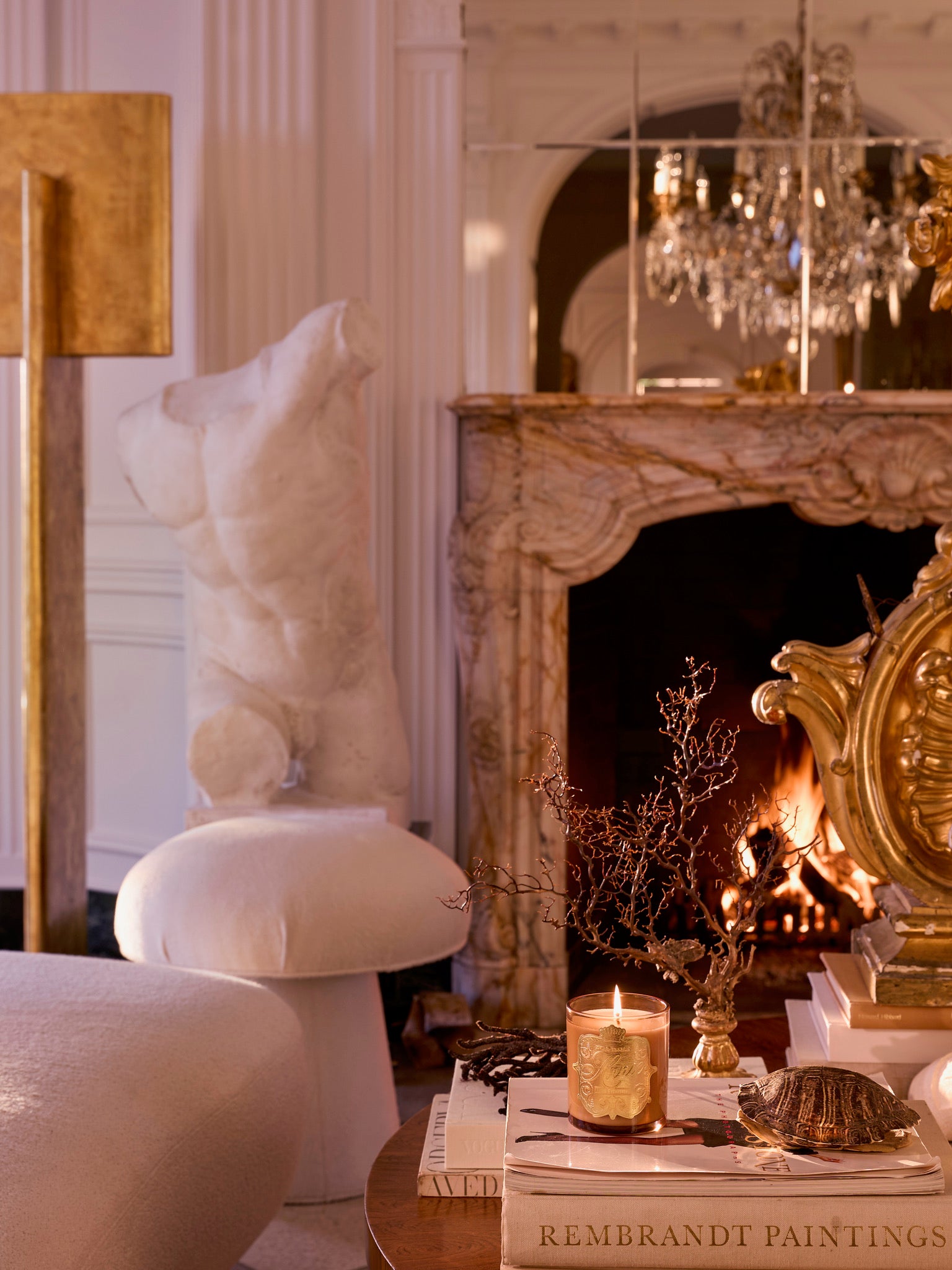 Cozy living room with a marble fireplace, glowing amber Apres Everything scented candle, books, turtle figurine, classical torso sculpture, and ornate decor. Crystal chandelier shimmers in the mirror above the mantel. Warm ambiance.