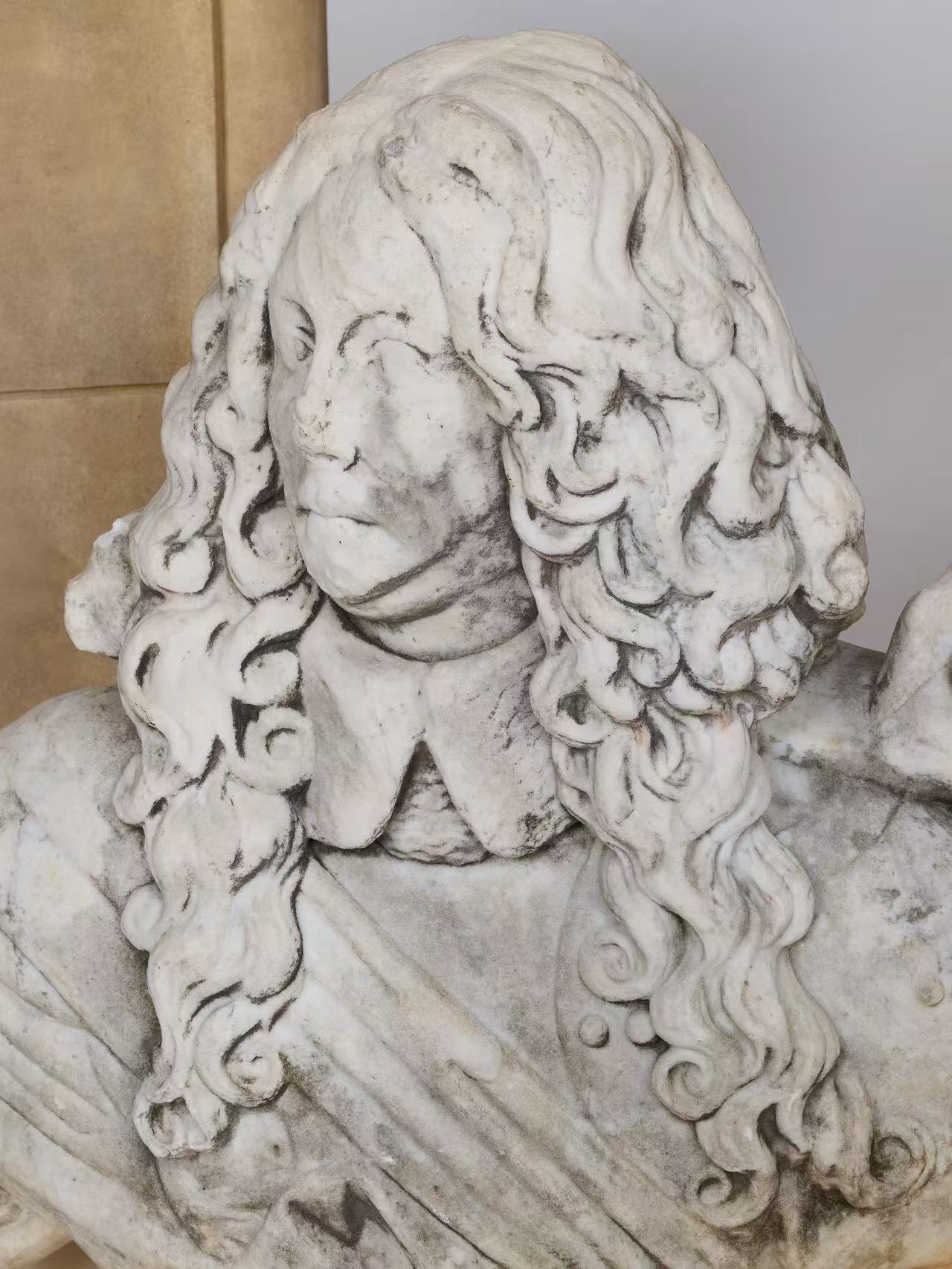 The Fragmentary Baroque-Style Stone Bust depicts a person with long, wavy hair in a collared garment. The facial features show wear and damage, and the bust is displayed against a smooth, light-colored background.