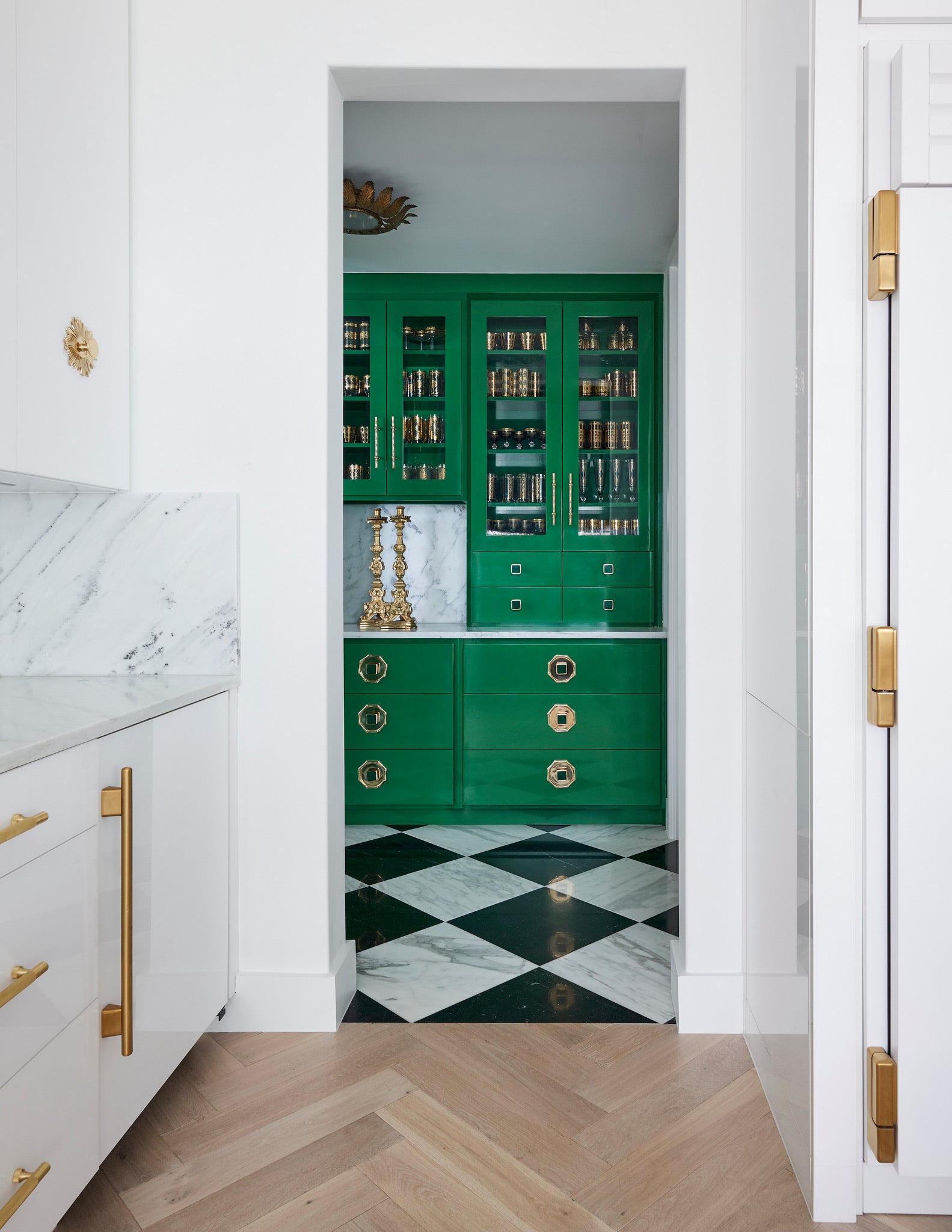 A small room with glossy emerald green cabinets, gold hardware, marble countertop, two gold candle holders, and black and white checkered floor, seen through a doorway from a white kitchen with brass accents and herringbone wood floor.