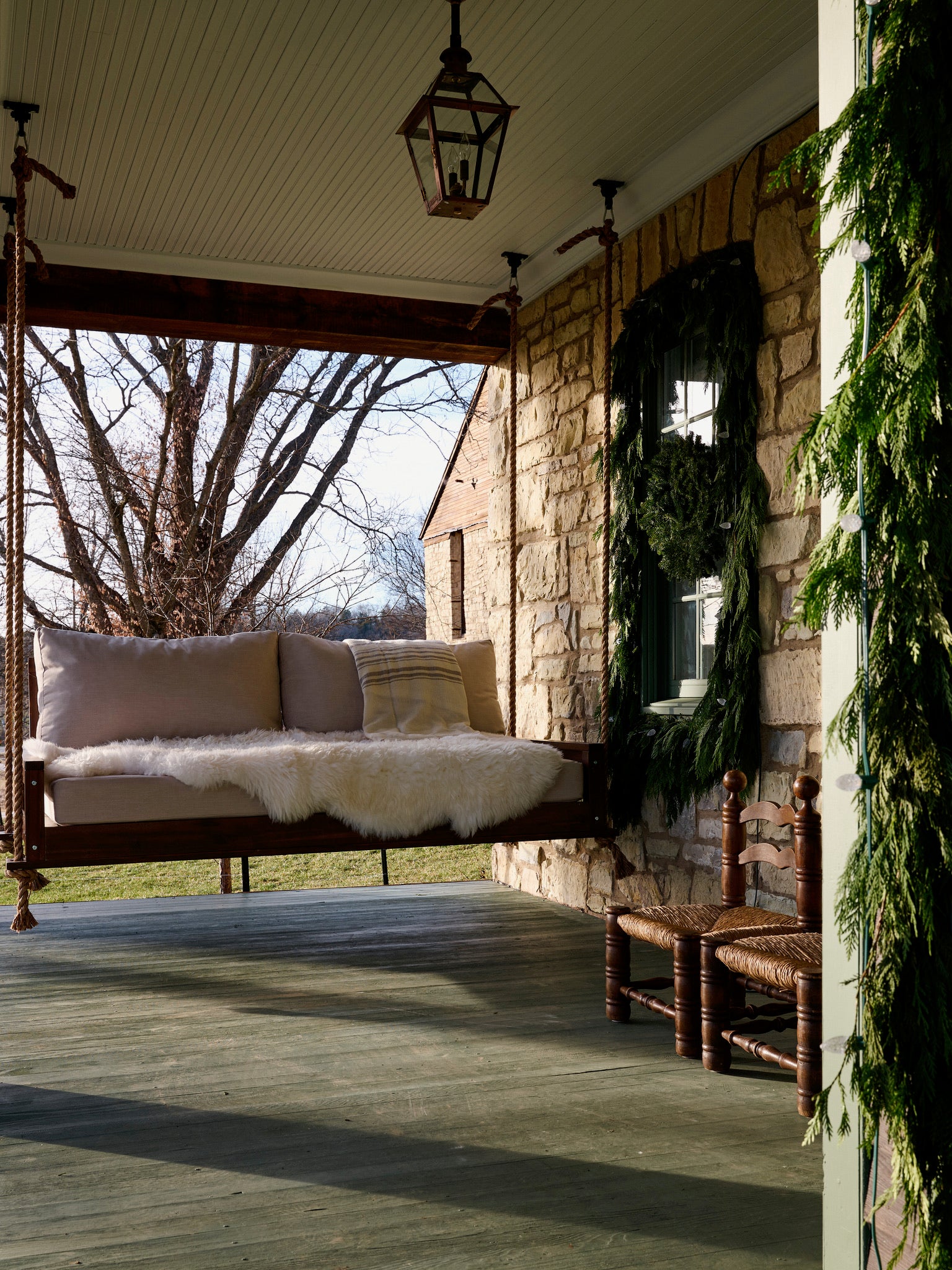 A cozy porch features a hanging daybed with cushions and a fur throw, rustic stone walls, a window with a wreath, wooden chairs, and greenery draped along a column, with bare trees visible outside.