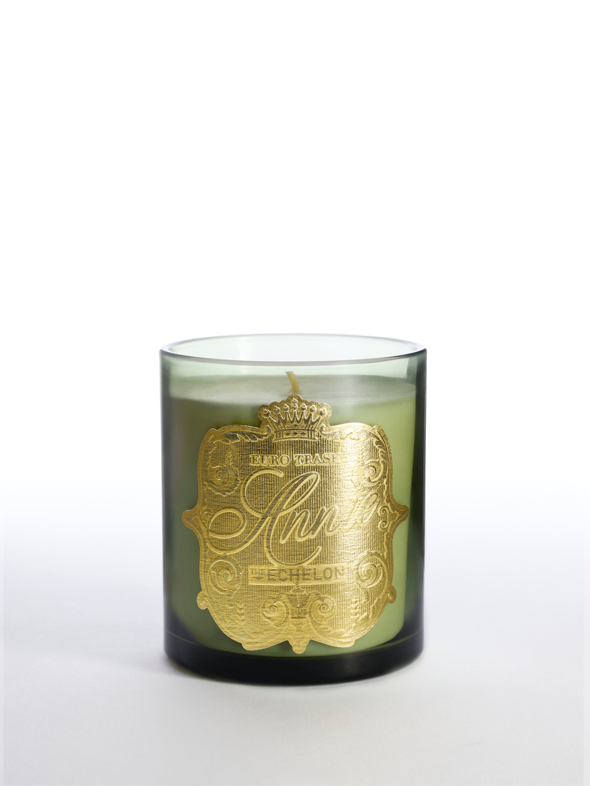 The Echelon is a green glass soy candle with a single wick, featuring an ornate gold label with decorative text and a crown design. It fills the air with warm spice notes and is shown against a plain white background.