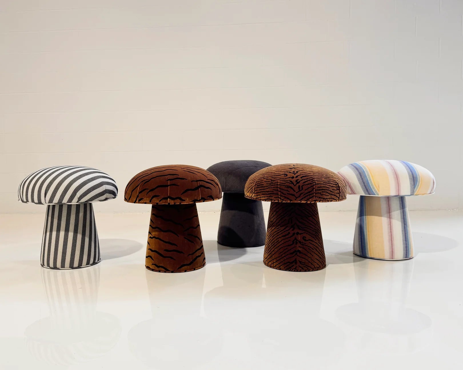 Five mushroom-shaped stools, including a handmade ottoman and The Forsyth Mushroom Pouf in Dedar 'A Tiger In The Orangerie' jacquard velvet, with stripes, tiger prints, and solids, are arranged on a shiny white floor against a plain wall.