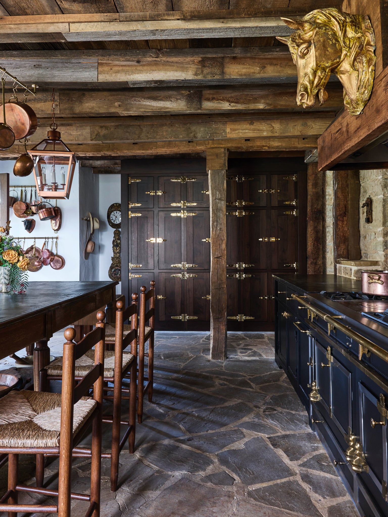 Rustic kitchen with exposed wood beams, stone floor, dark wood cabinets with brass hardware, a black stove, wooden chairs, and a gold horse head sculpture mounted on the wall. Copper pots hang from a rack.
