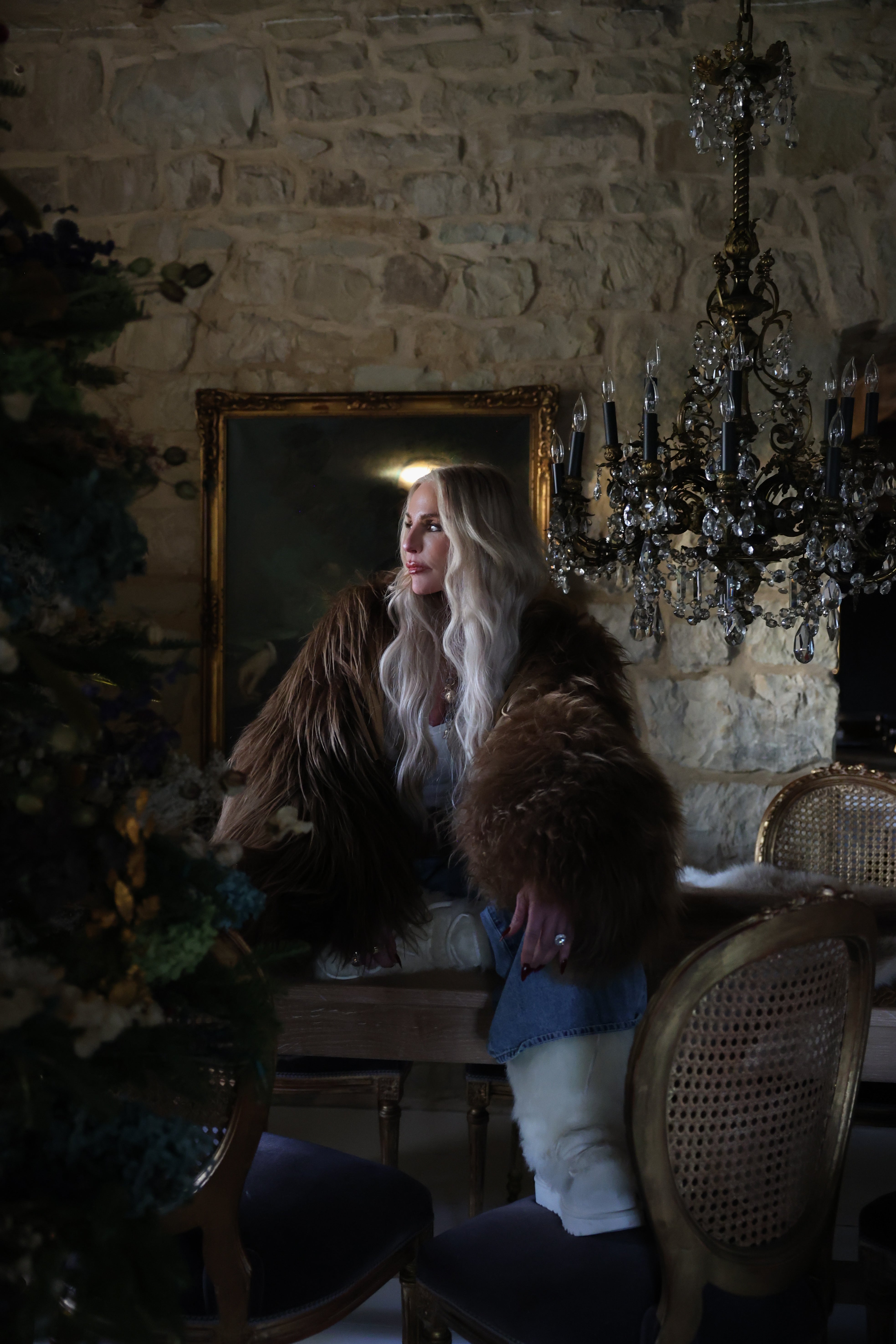 A woman with long blonde hair, wearing a large brown fur coat, sits at a wooden table in a rustic room with stone walls, ornate chairs, and a chandelier hanging above her. A dark painting and foliage are in the background.