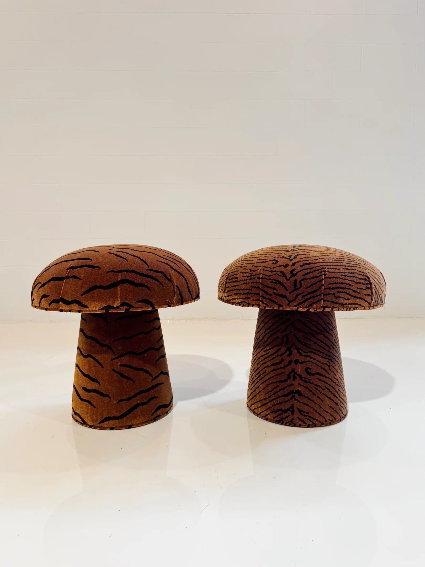 Two Forsyth Mushroom Poufs in Dedar 'This Is The Tiger Speaking' upholstery, featuring brown and black tiger stripes, rest on a white floor against a plain, light-colored wall.