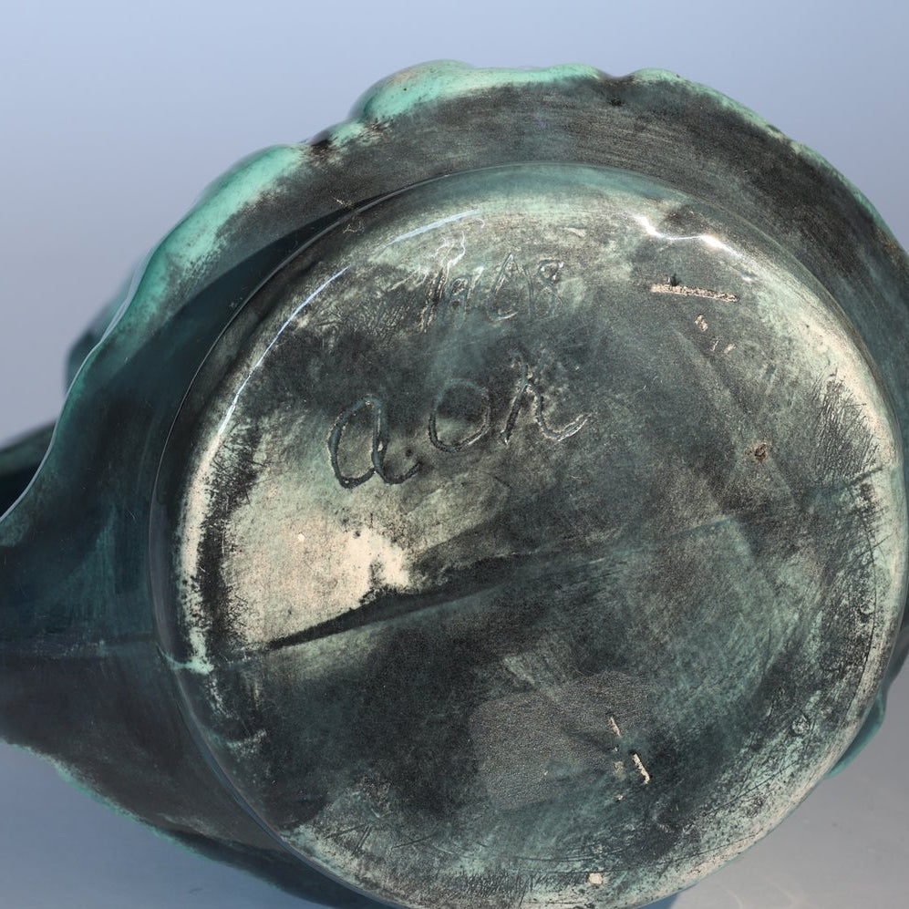 The base of this Vintage Green Majolica Style Grape Motif Ceramic Pitcher by Euro Trash has a greenish patina, hand-etched markings like “AOK” and possibly a date on the underside, plus surface wear and discoloration.