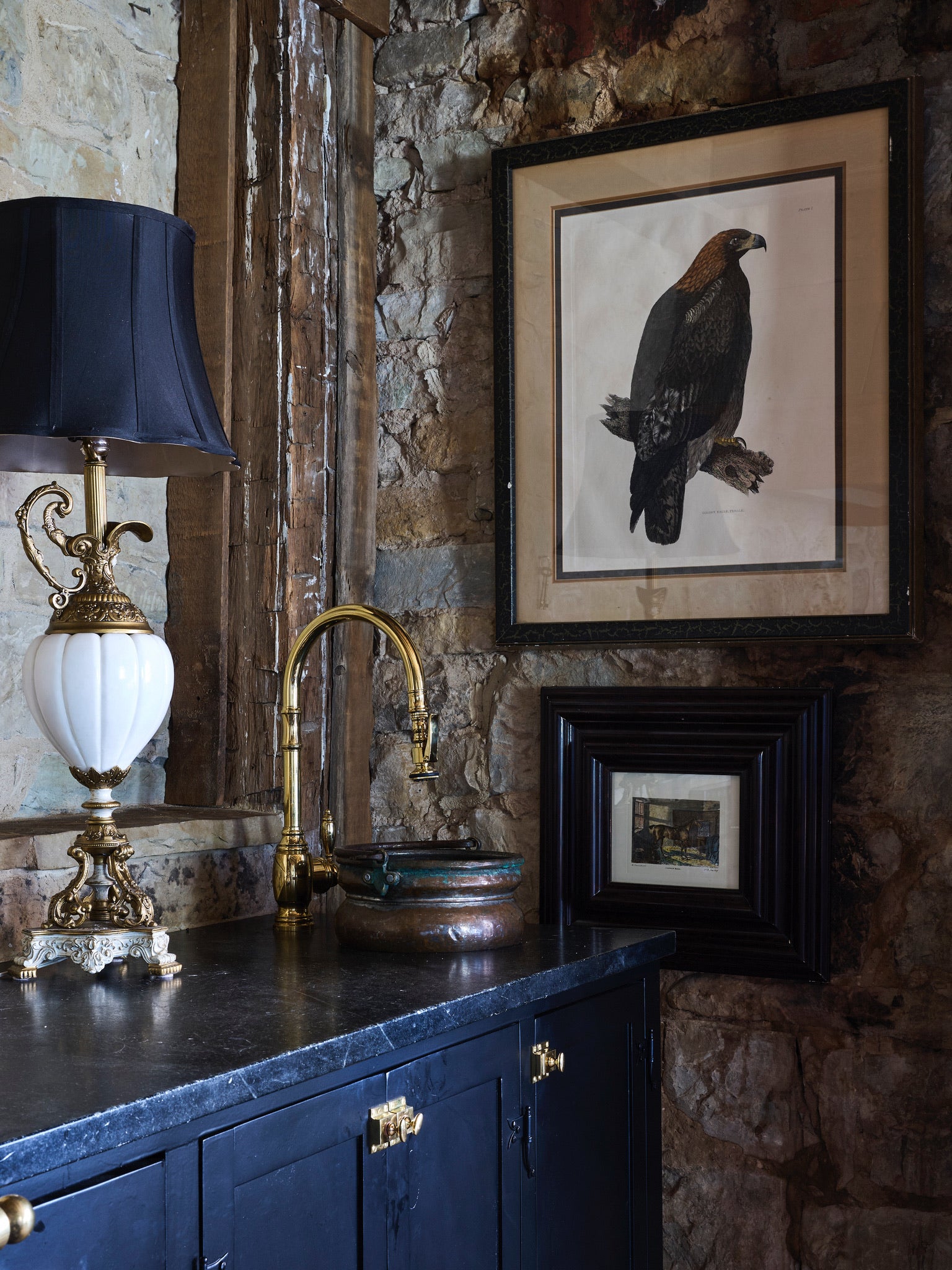 Stone-walled room with a black cabinet, ornate gold lamp, brass faucet, and two framed artworks on the wall—one featuring a large bird, the other a small landscape.