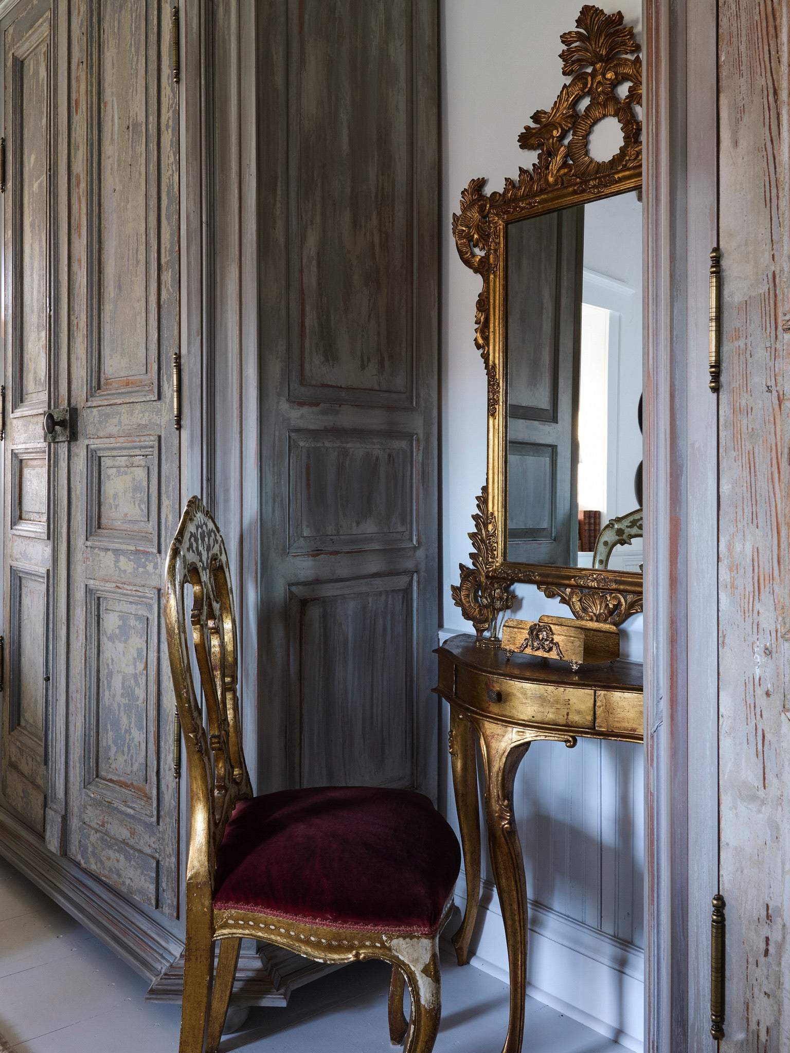 An ornate gold vanity table with a matching mirror and a red velvet chair sits next to distressed wooden wardrobe doors in a vintage-style room.