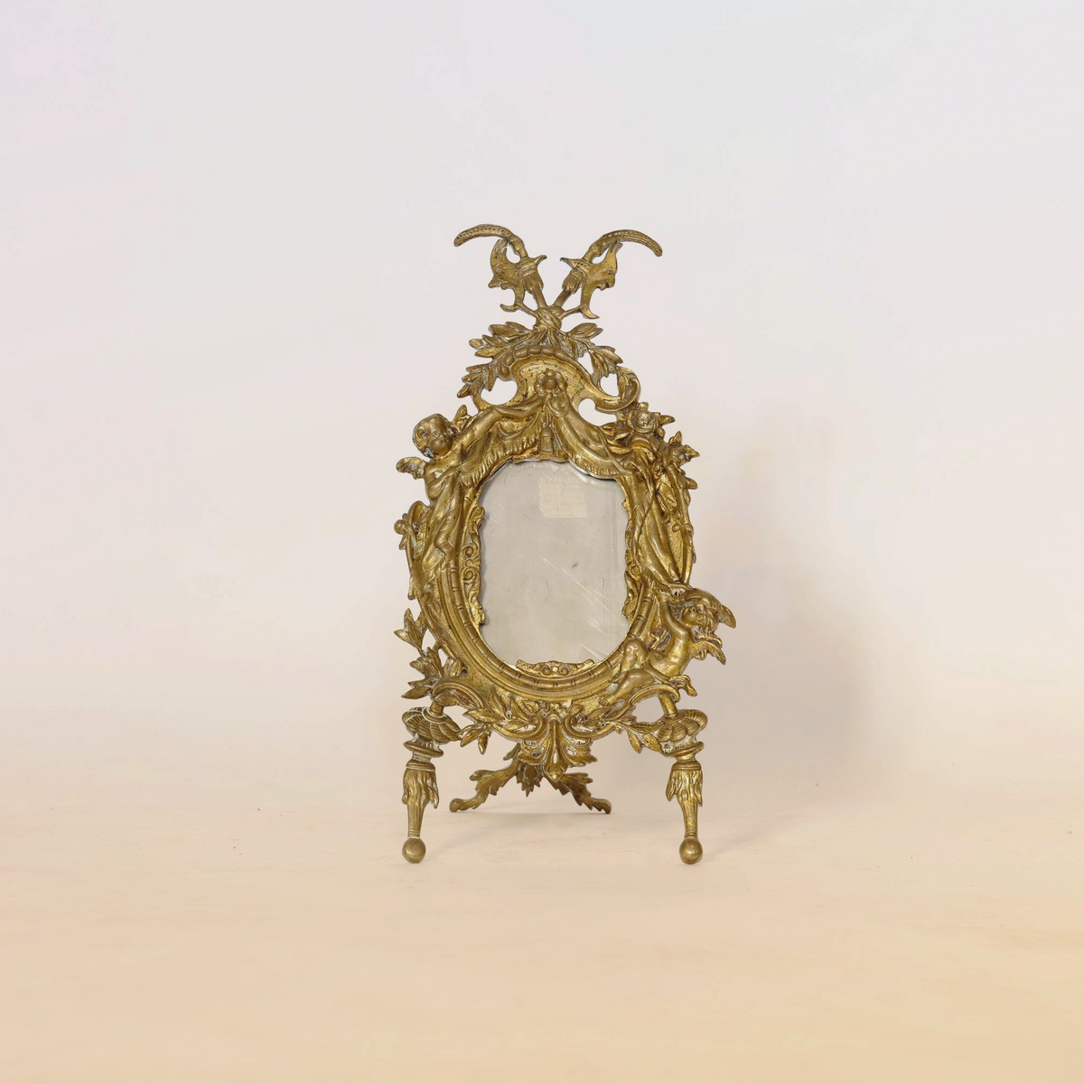 The Antique Gilt Bronze Rococo Easel Mirror by Euro Trash, featuring intricate detailing and ornate Rococo ornamentation, stands upright on three legs against a plain, light background.