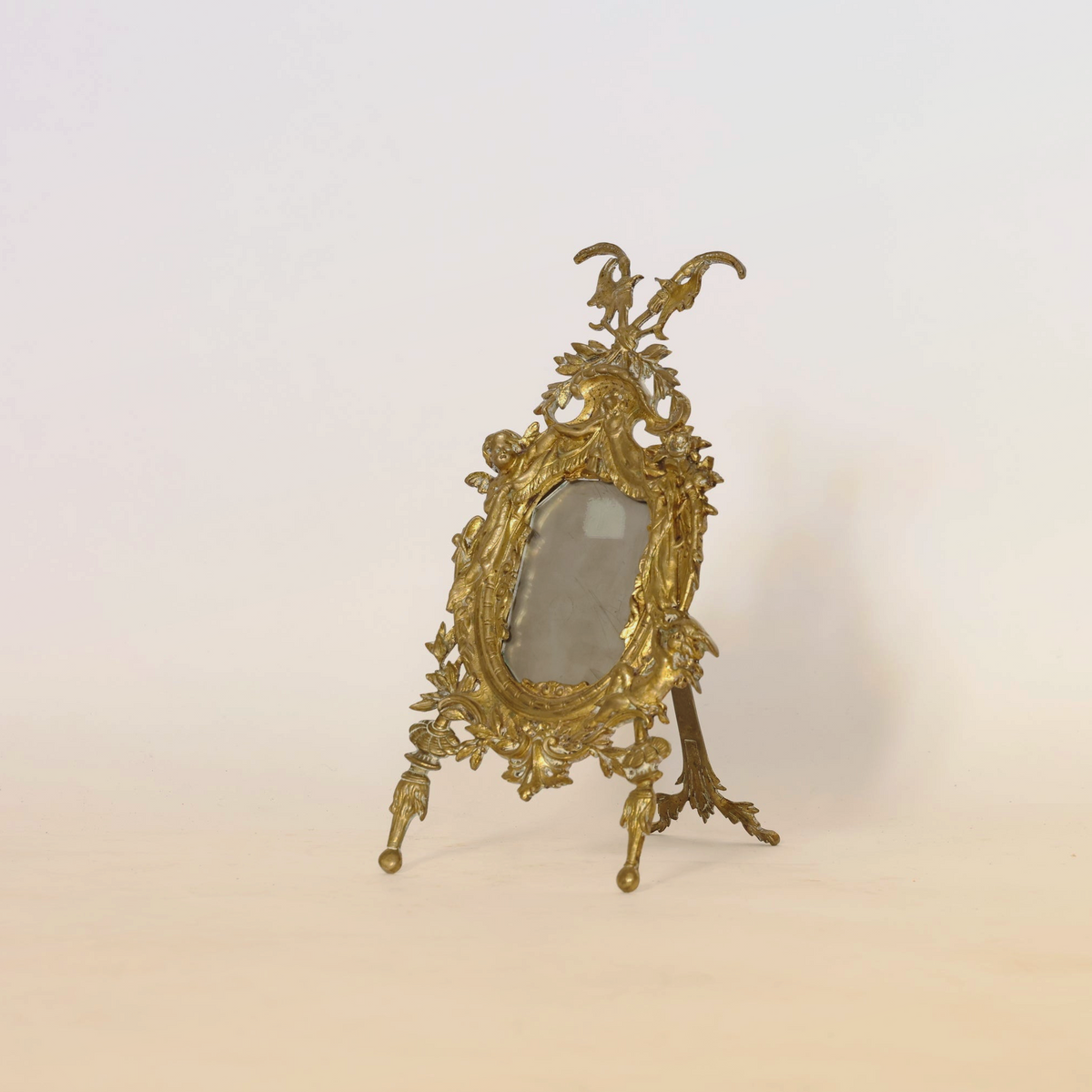 The Antique Gilt Bronze Rococo Easel Mirror by Euro Trash features an ornate, oval gold frame adorned with intricate floral motifs and classic Rococo detailing. Standing elegantly on three legs against a simple, light backdrop, the frame evokes antique charm and is fitted with a piece of cloudy glass.