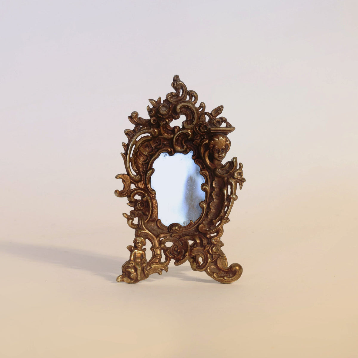 The Antique Gilt Bronze Rococo Table Mirror by Euro Trash Annie features an ornate, vintage-style design with an intricately crafted gilt bronze frame adorned with decorative Rococo scrolls and small sculpted figures.
