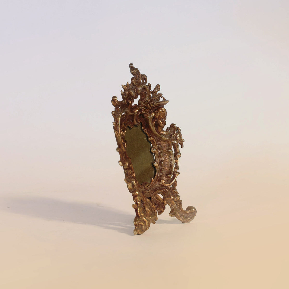 An ornate Euro Trash Annie Antique Gilt Bronze Rococo Table Mirror with intricate gold detailing stands angled on a pale surface against a light background, evoking the elegance of vintage design.
