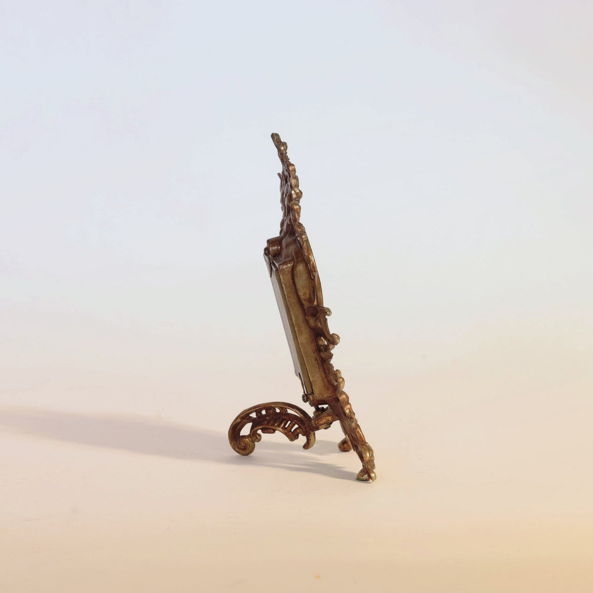 The Antique Gilt Bronze Rococo Table Mirror by Euro Trash Annie is displayed from a side view, standing upright on a simple pale background. Reminiscent of Rococo design, the mirror features ornate detailing and a gracefully curved support leg.