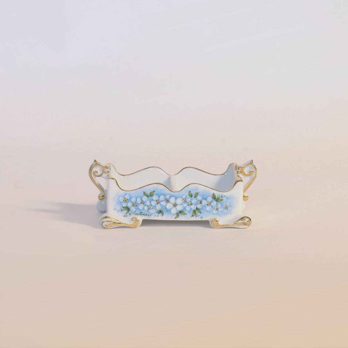 The Antique Hand-Painted Porcelain Floral Cachepot by Euro Trash Annie is a small, ornate rectangular porcelain piece featuring gold trim and legs, adorned with hand-painted blue and white floral motifs on the front set against a pale background.