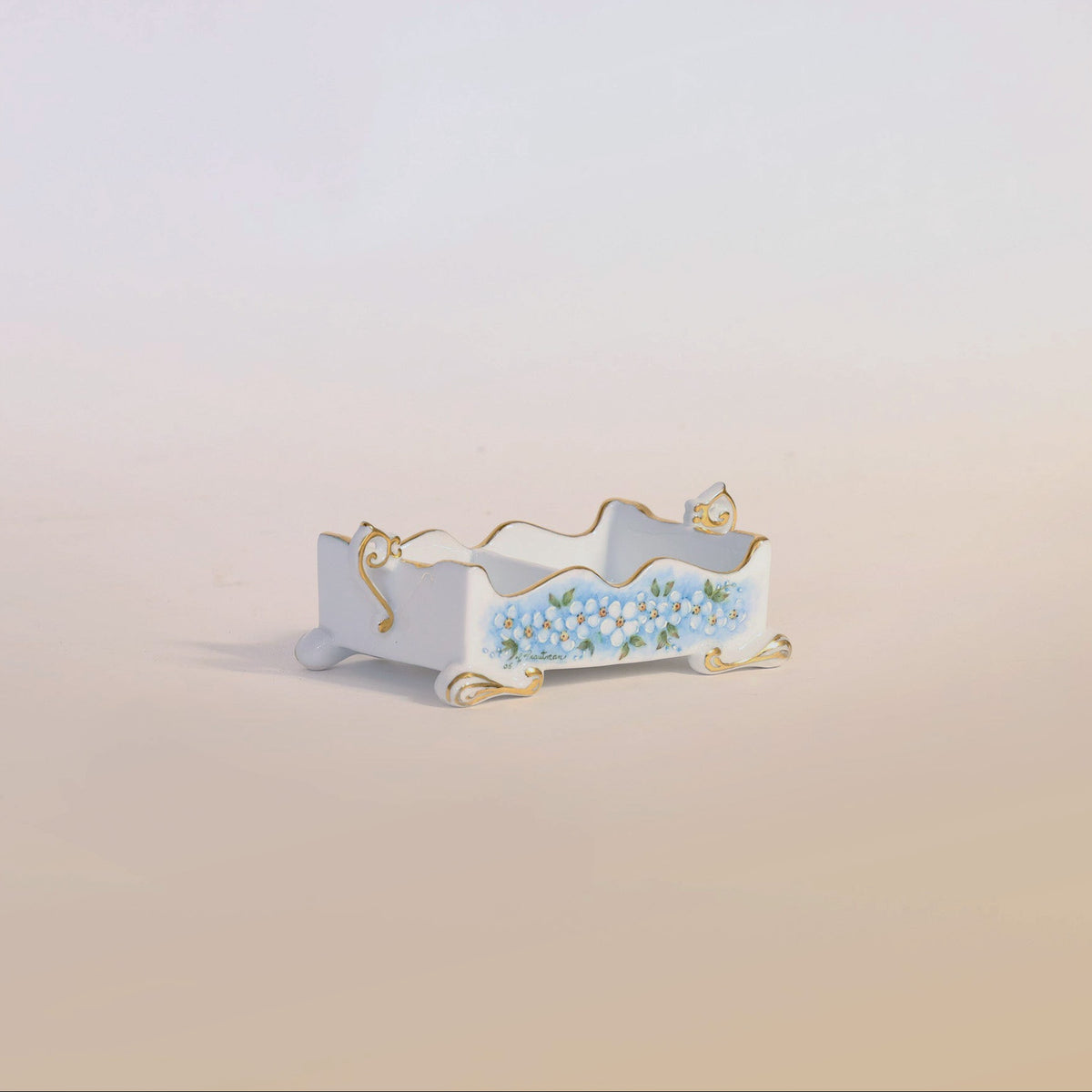The Antique Hand-Painted Porcelain Floral Cachepot by Euro Trash Annie features a compact, rectangular design with ornate feet and elegant gold trim, showcasing intricate blue and white floral motifs on fine porcelain, beautifully displayed against a soft, light background.