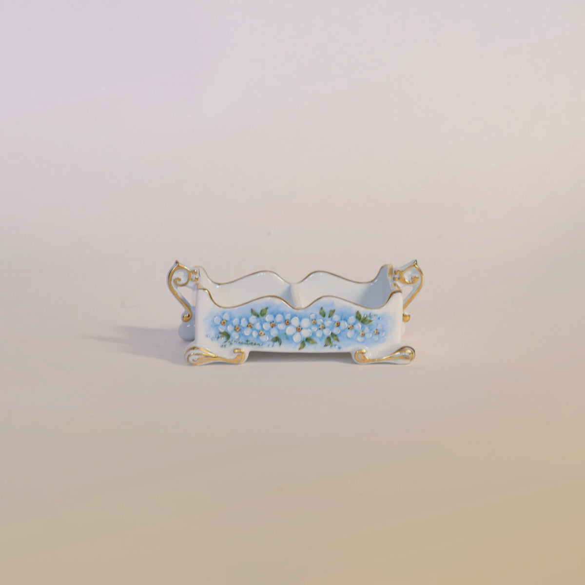 The Antique Hand-Painted Porcelain Floral Cachepot by Euro Trash Annie features a small, rectangular silhouette adorned with gold accents and delicate blue floral motifs, all hand-painted along its sides against a simple, light backdrop.
