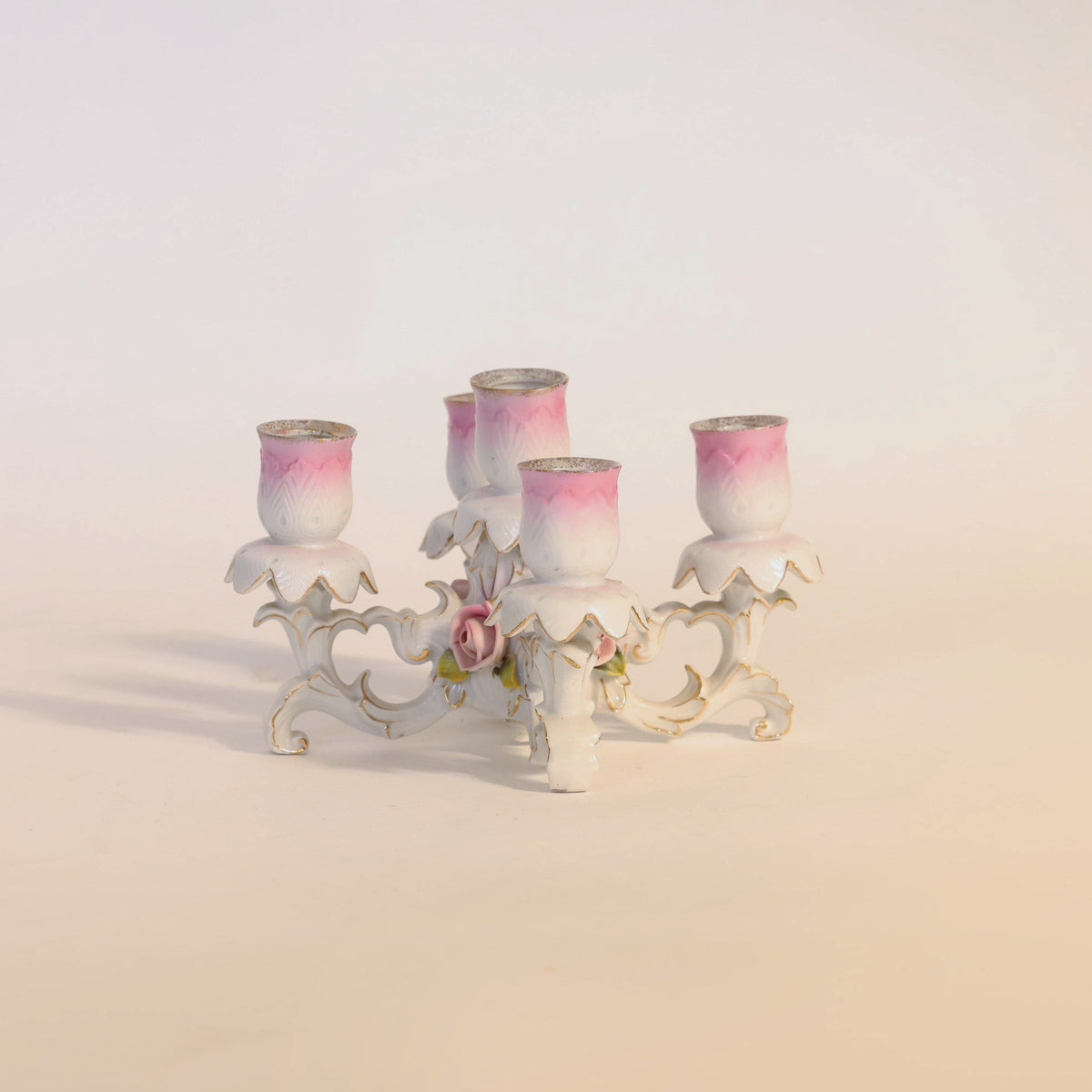 The Antique Porcelain Floral Candelabra by Euro Trash Annie features four candle holders shaped like pink and white tulips, exquisite floral detailing, and a delicately curved base reminiscent of classic antique porcelain designs.