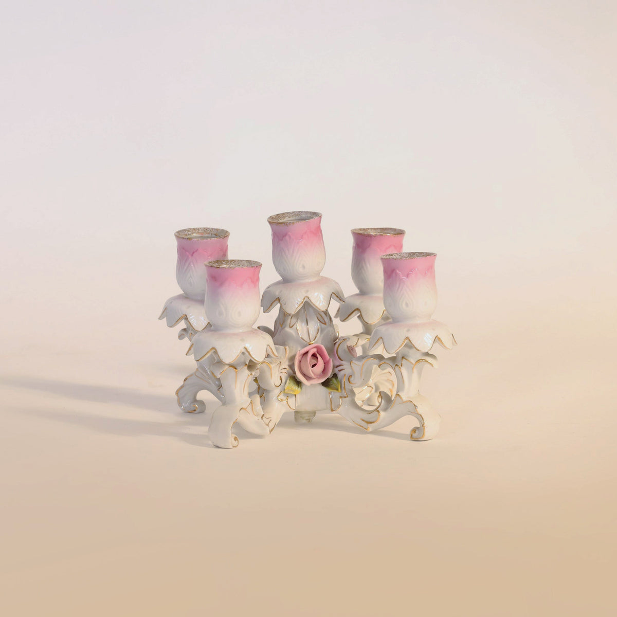 The Antique Porcelain Floral Candelabra by Euro Trash Annie is a decorative white ceramic candle holder featuring five tulip-shaped pink and white holders, an ornate leaf-inspired base, and a central ceramic rose accent. Evoking the timeless elegance of antique porcelain, this floral candelabra makes a charming centerpiece for any space.