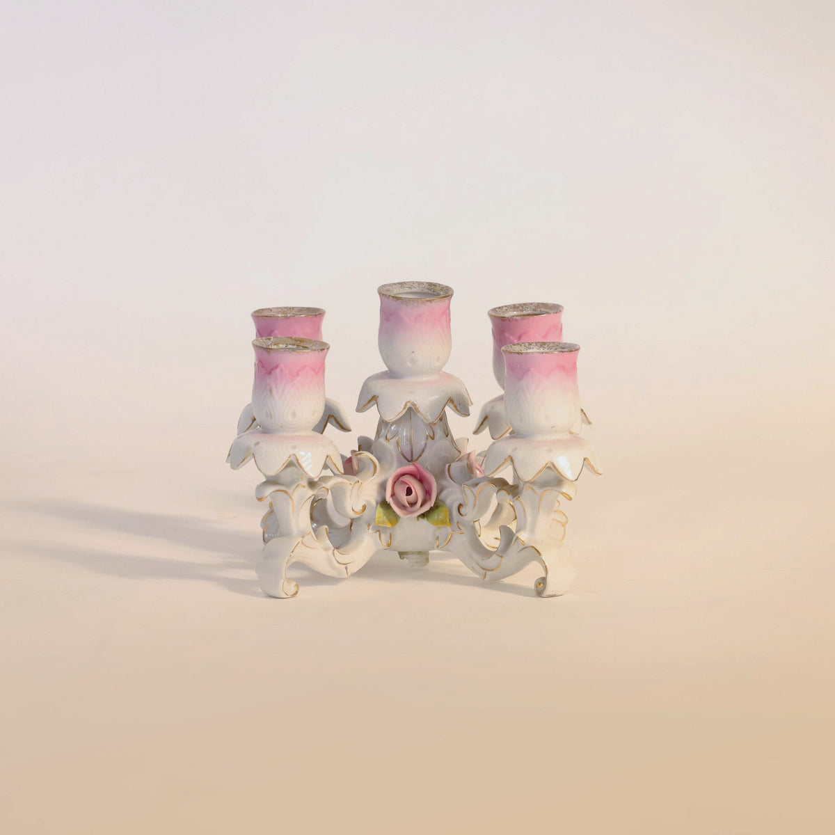 The Antique Porcelain Floral Candelabra by Euro Trash Annie is a vintage decorative candle holder crafted from antique porcelain, featuring four pink holders with gold trim and ornate white detailing. Its centerpiece is beautifully adorned with a delicate pink rose, enhancing the timeless elegance of this piece.