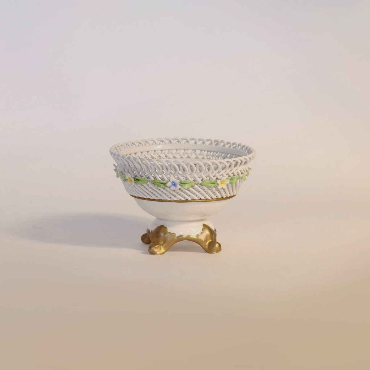 The Antique Porcelain Reticulated Basket Compote by Euro Trash Annie features a white bowl with an intricate lattice design and delicate pierced detailing, adorned with small blue and yellow flowers. It rests atop a gold-colored, ornate base against a plain, light background—an exquisite piece that brings timeless charm to any setting.
