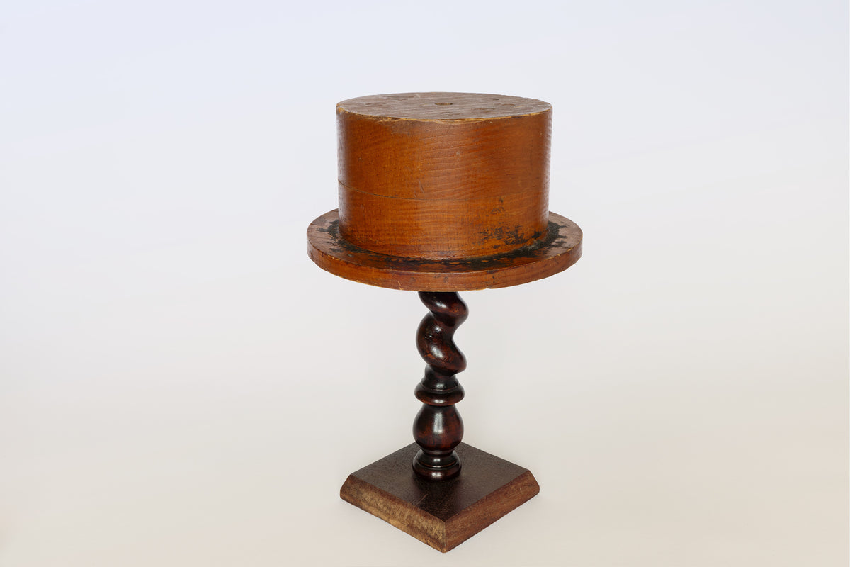 An Antique Wooden Hat Mold by Euro Trash is displayed on a dark, twisted pedestal stand with a square base, forming an eye-catching sculptural piece set against a plain white background.
