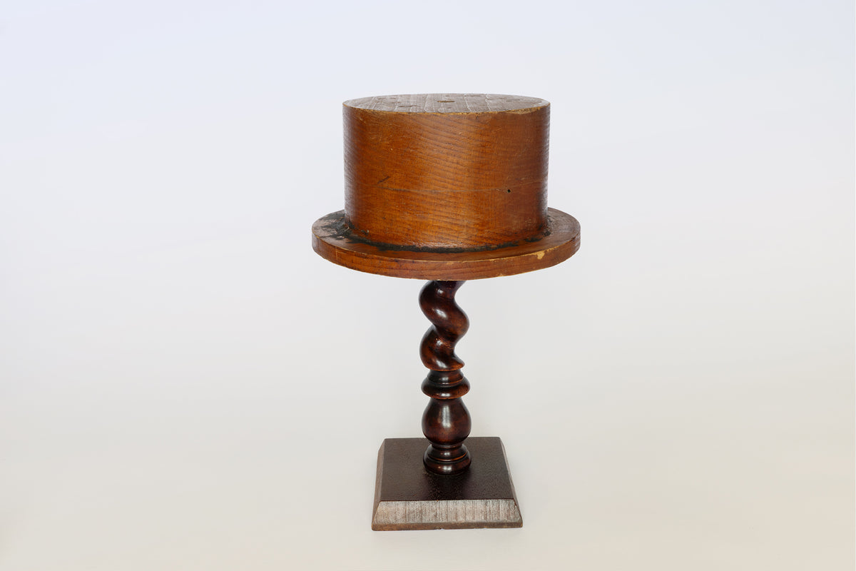 The Euro Trash Antique Wooden Hat Mold features a cylindrical top, elegantly perched on a twisted pedestal with a square base. Its sculptural design not only serves as an ideal tool for shaping hats but also stands out as a unique decorative accent, shown here against a plain white background.