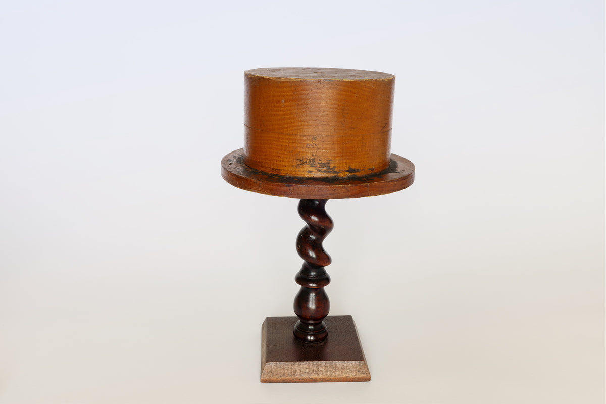 A sculptural object, the Euro Trash Antique Wooden Hat Mold features a cylindrical top and wide brim, set atop a dark, twisted wooden stand with a square base against a plain white background—perfect as a decorative interior piece.