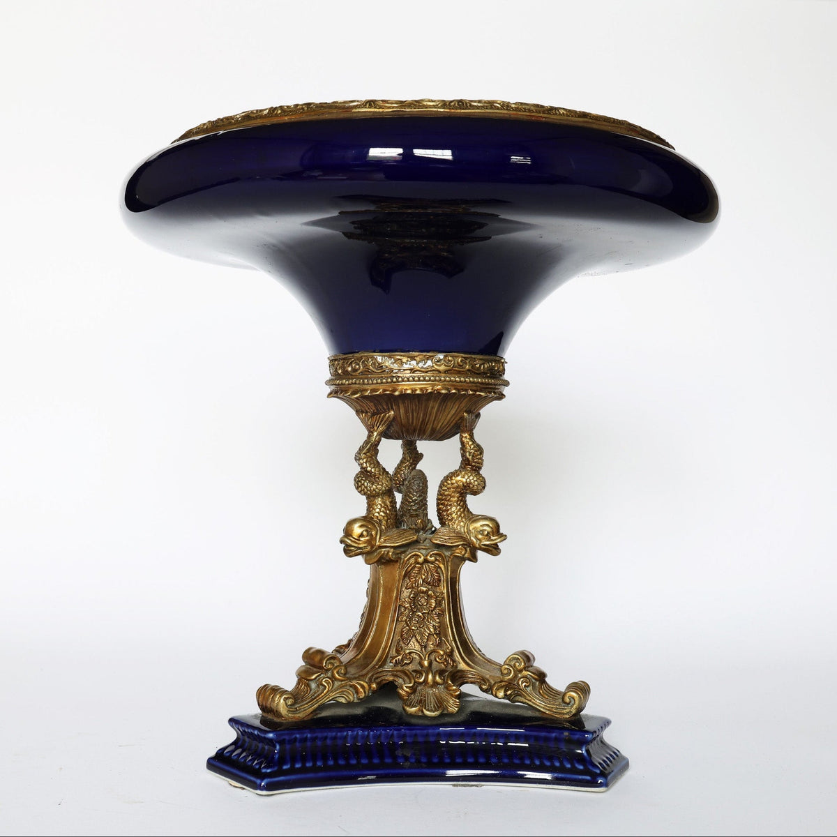 Euro Trash Antique Cobalt Porcelain & Gilt Bronze Centerpiece Tazza features a glossy deep blue top, ornate gilt bronze accents, and a base with sculpted gold dolphins and scrollwork, set against a plain white background.