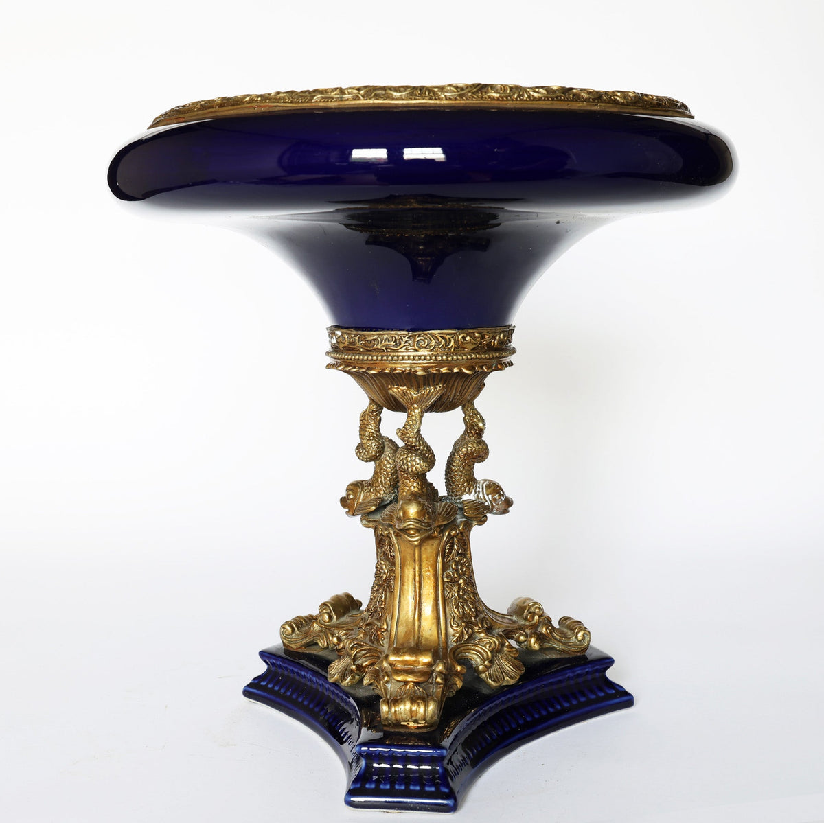 The Euro Trash Antique Cobalt Porcelain & Gilt Bronze Centerpiece Tazza features a glossy cobalt bowl top, ornate gilt bronze details, sculpted gold base with figures and patterns, and stands on a blue four-cornered platform.