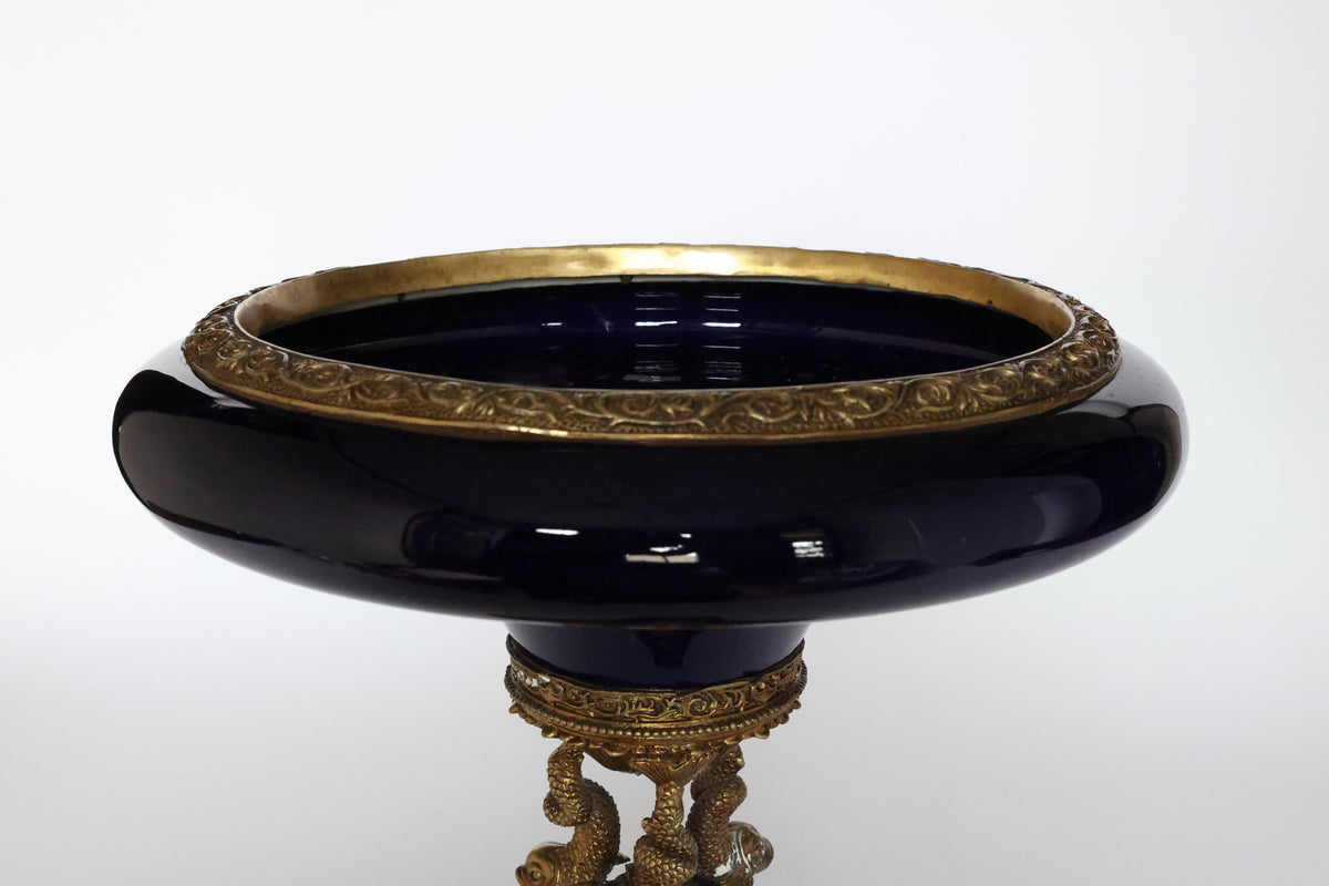The Antique Cobalt Porcelain & Gilt Bronze Centerpiece Tazza by Euro Trash features a deep blue round bowl with an ornate gold rim, set on a decorative gilt bronze stand with intricate designs and sculpted figures against a plain white background.