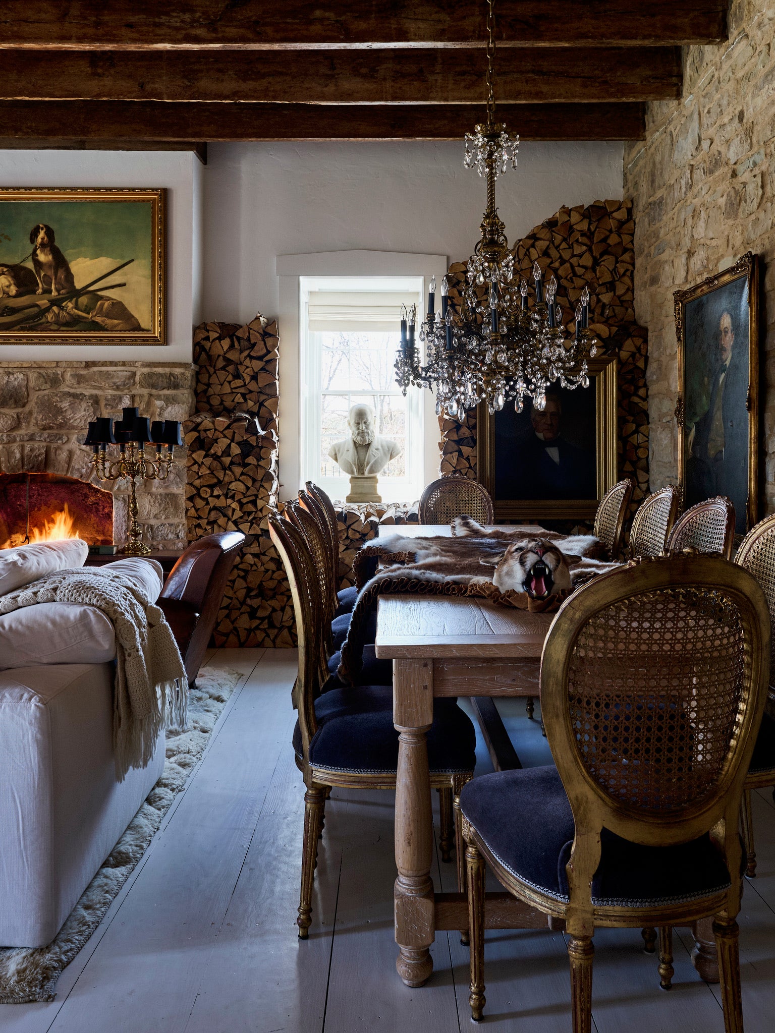 A rustic dining room with a wooden table, upholstered chairs, stone walls, a chandelier, a fireplace, and stacks of firewood. Portraits and a bust statue decorate the space, with a window letting in natural light.