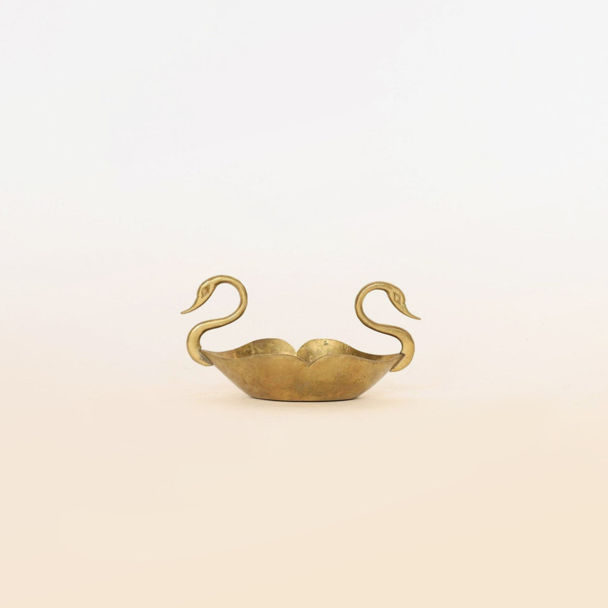 The Brass Swan Planter by Euro Trash Annie is a sculptural brass bowl featuring two swan-shaped handles facing opposite directions, making it a striking planter and decorative accent, beautifully set against a pale beige background.