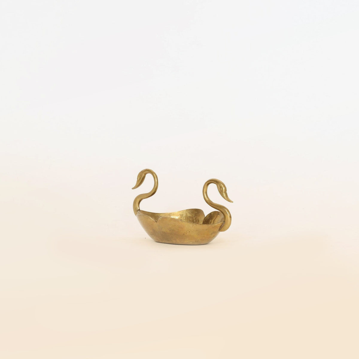 The Brass Swan Planter by Euro Trash Annie is a small, golden decorative bowl featuring two handles sculpted as graceful swan heads and necks; this elegant piece serves as a refined sculptural accent, beautifully set against a plain, light background.