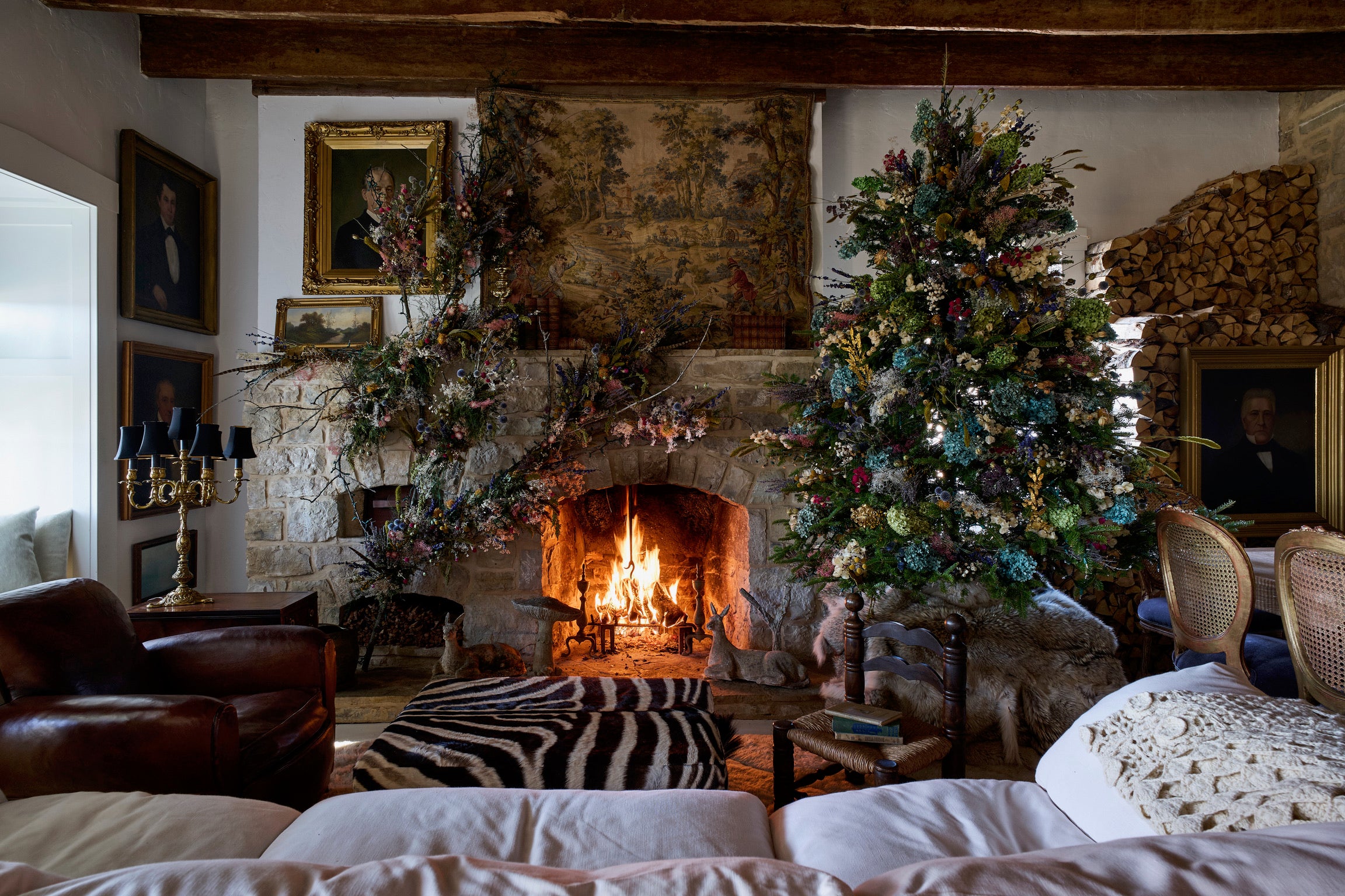 A cozy living room with a roaring fireplace, decorated with two Christmas trees, floral garlands, and framed portraits. Wooden beams, stacked firewood, and comfy seating add to the rustic, festive atmosphere.